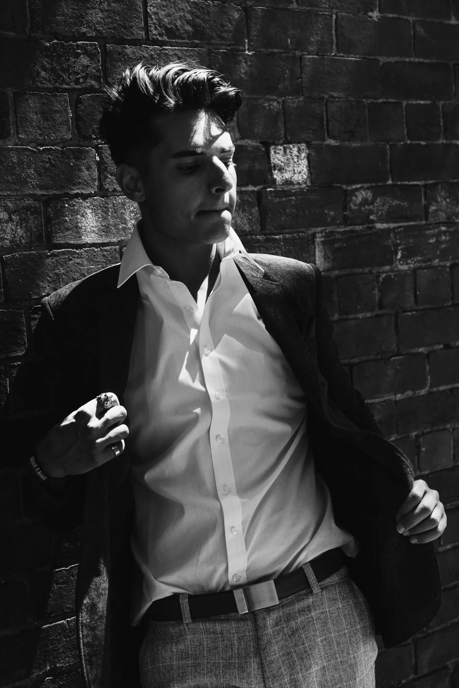 A young man in a white dress shirt and dark blazer, leaning against a brick wall, with shadows cast across his face, in black and white.