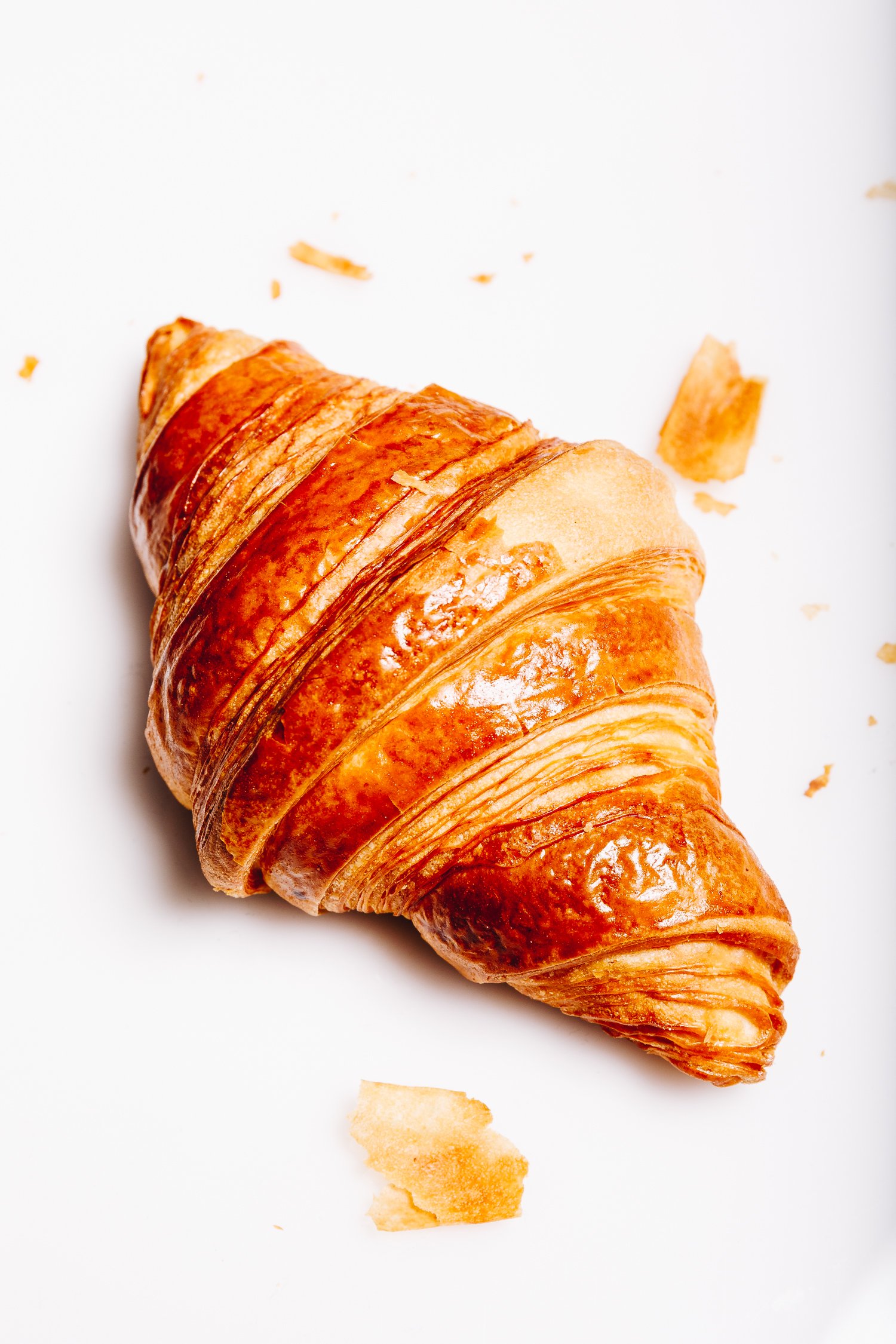 A golden-brown croissant on a white surface with small crumbs around it.