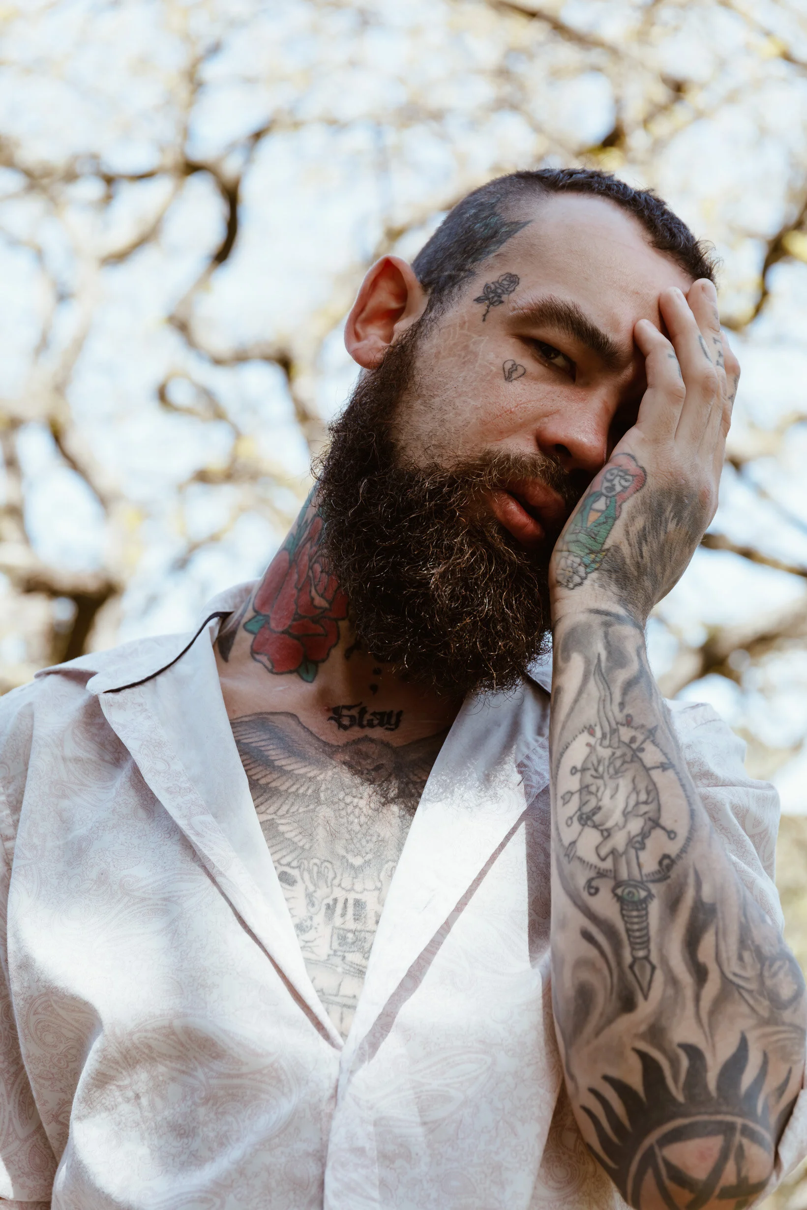 A bearded man with tattoos covering his neck and arms, wearing a light-colored patterned shirt, holds his face with one hand, standing outdoors with a background of trees.