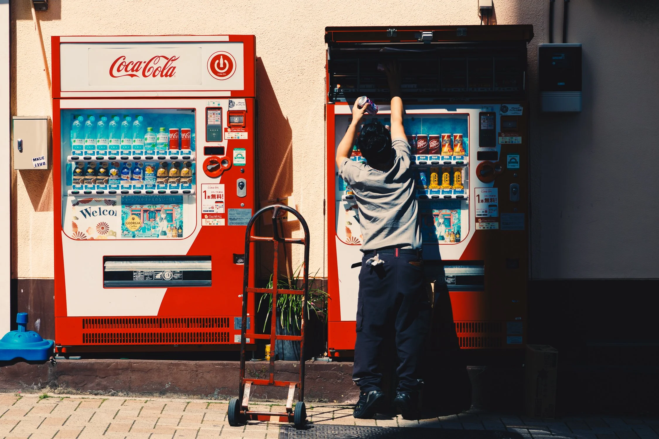 A man wearing a gray shirt and black pants is using a vending machine to buy drinks outside a building. There are two vending machines side by side, and the man is reaching into the top row of the right vending machine. A hand truck is positioned in 