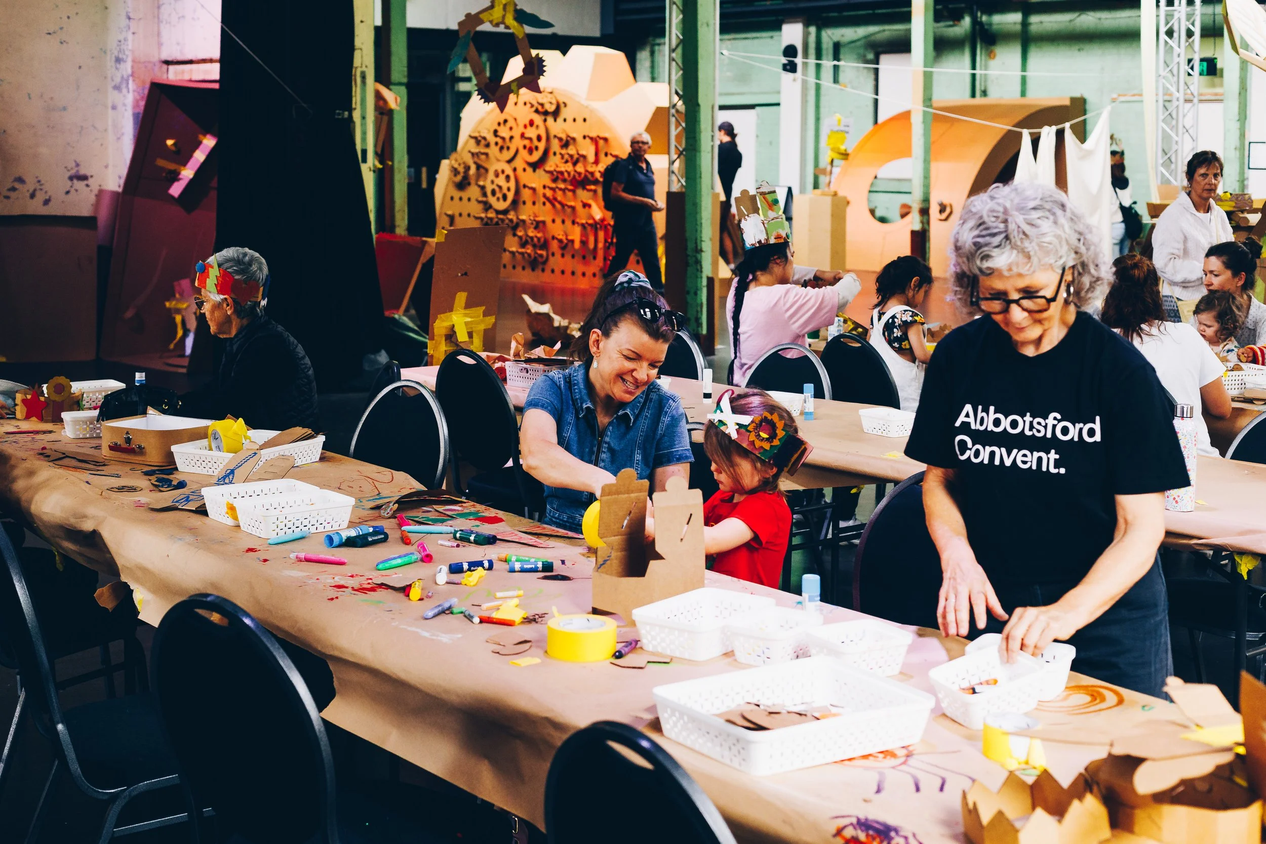 People engaging in arts and crafts activities at a long table covered with brown paper in an artist workshop or community space.