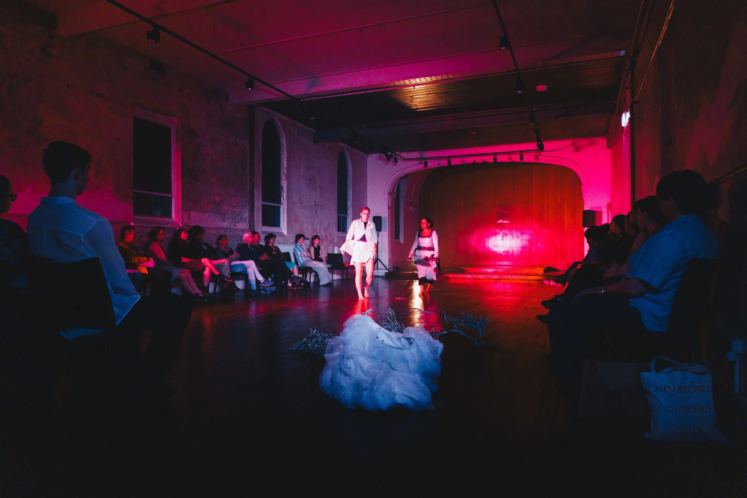 Two performers are on stage in a dimly lit room with pink and purple lighting, surrounded by an audience seated along the sides, watching the performance.