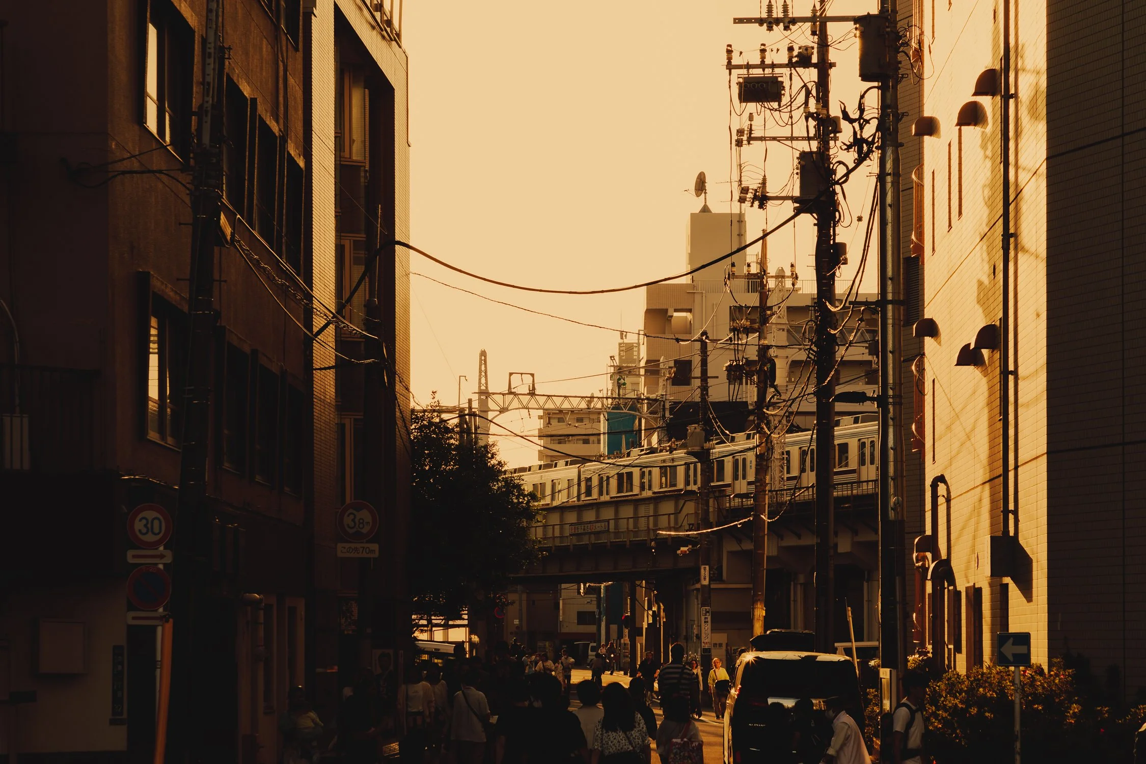 City street scene at sunset with people walking, buildings, utility poles, and a train in the background.