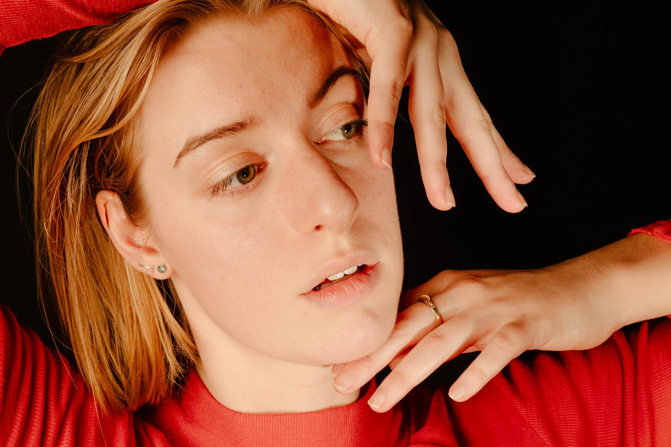 A close-up portrait of a young woman with red hair wearing a red top, posing with her hand near her face, against a black background.