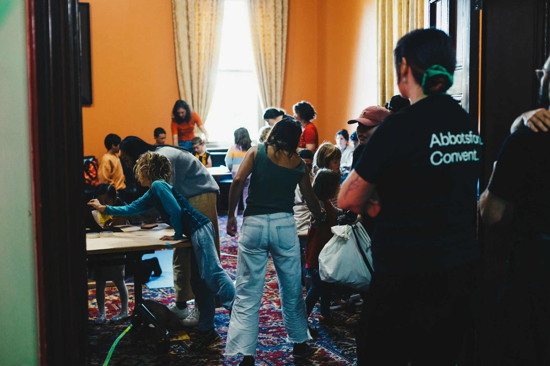 Children and adults participating in a group activity or workshop in a room with orange walls, large windows, and patterned carpet.