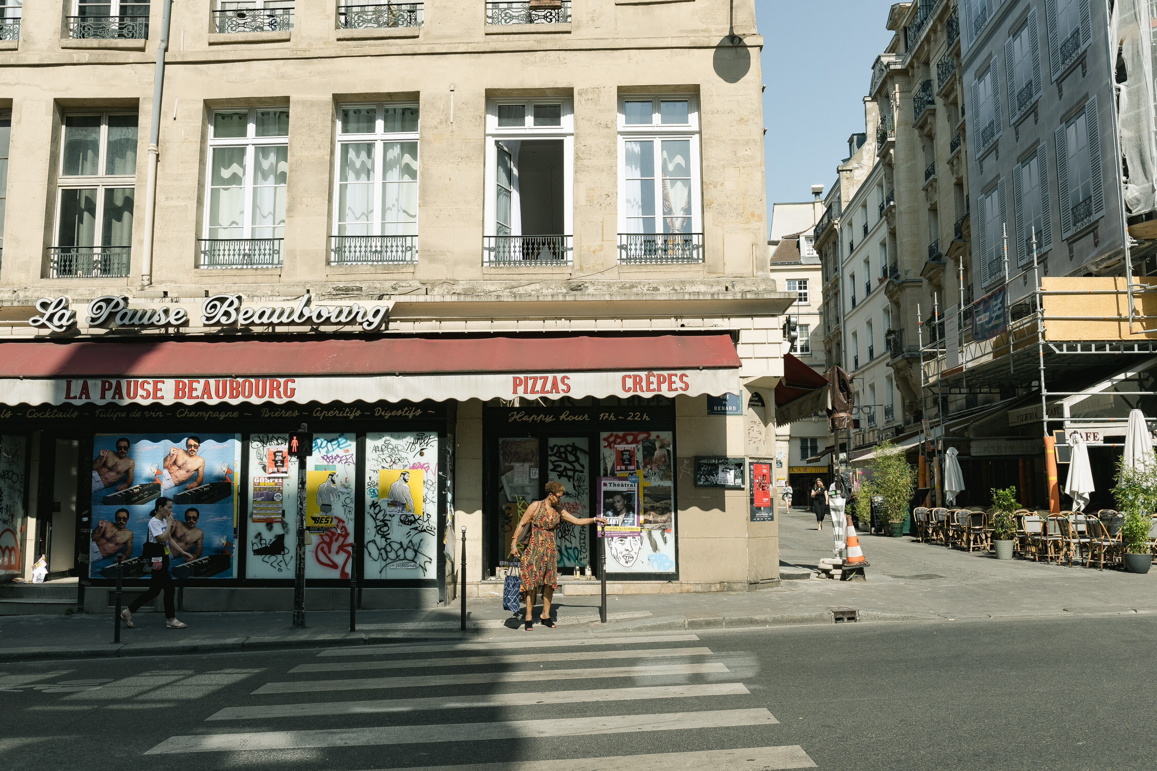 Street scene in front of a restaurant called La Pause Beaubourg, featuring graffiti-covered windows, a woman at a crosswalk, and outdoor seating area with tables and umbrellas on the sidewalk.