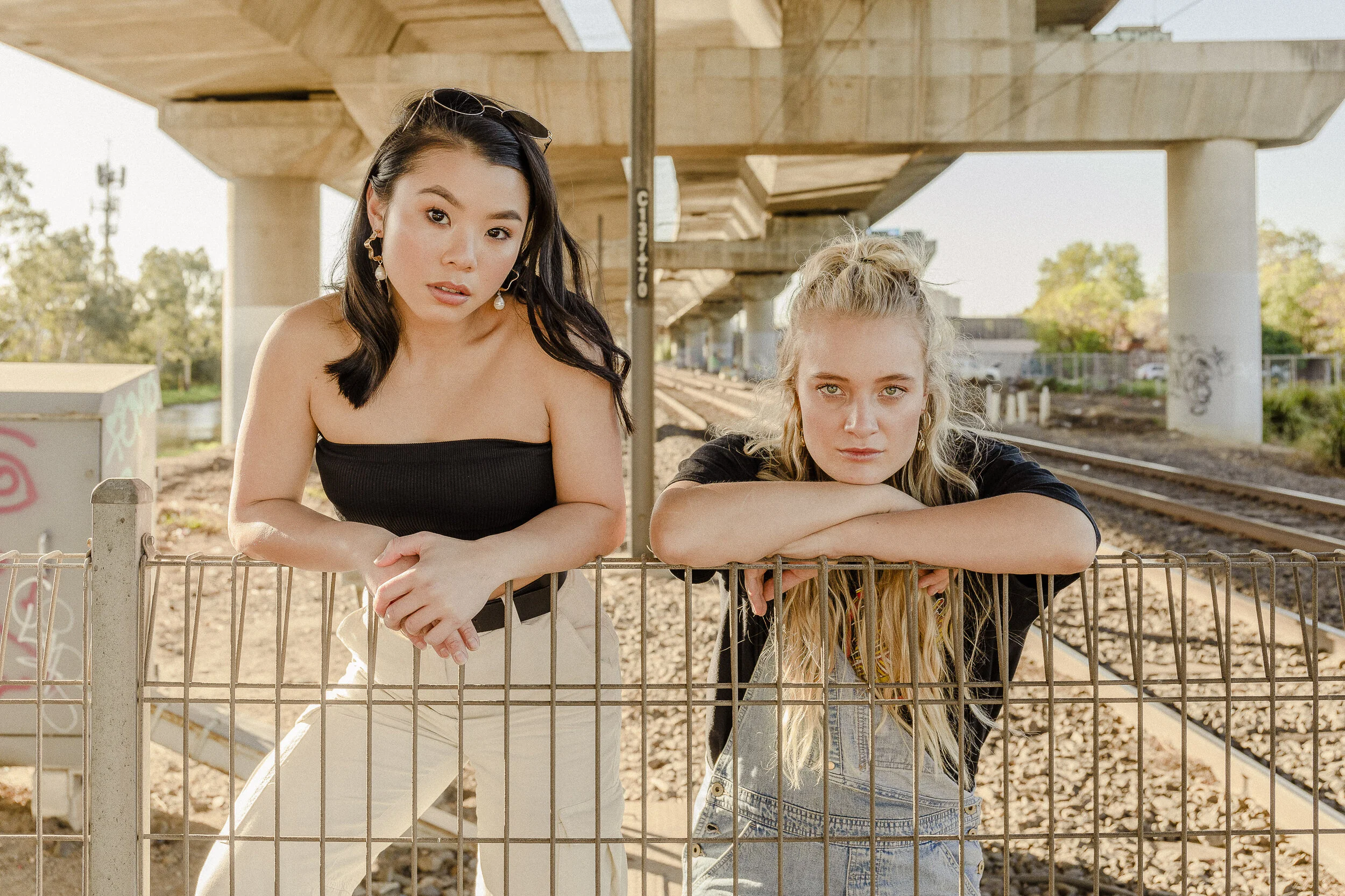 Two young women leaning on a rusty metal fence at a train track under a bridge. One has dark hair, wearing a black strapless top and beige pants, the other has blonde hair, wearing a black shirt and denim overalls. The scene is outdoors during daylig
