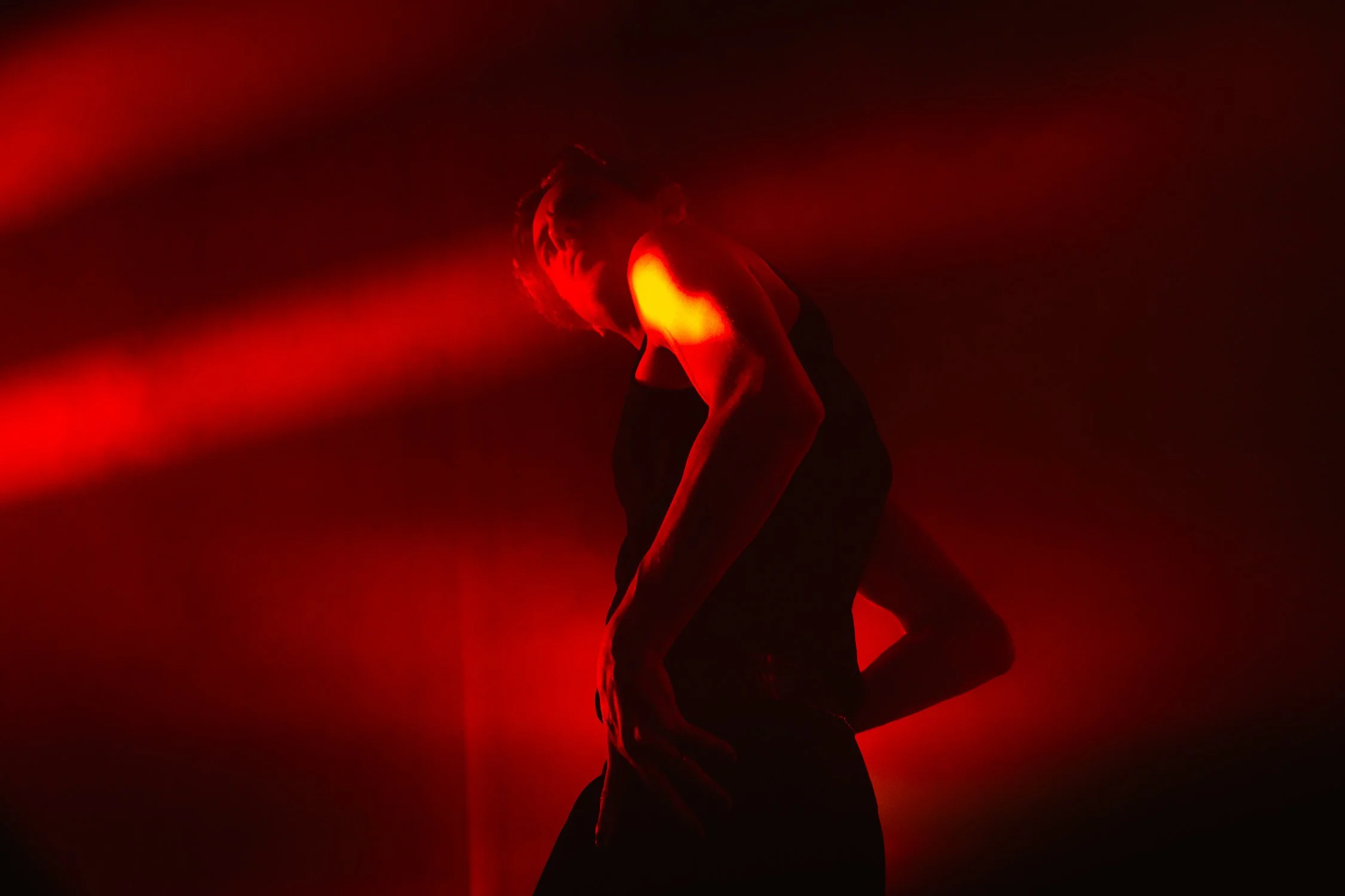 A person stands in a dark space illuminated by red stage lighting, with their head tilted down and hand on their thigh, creating a silhouette against the red glow.