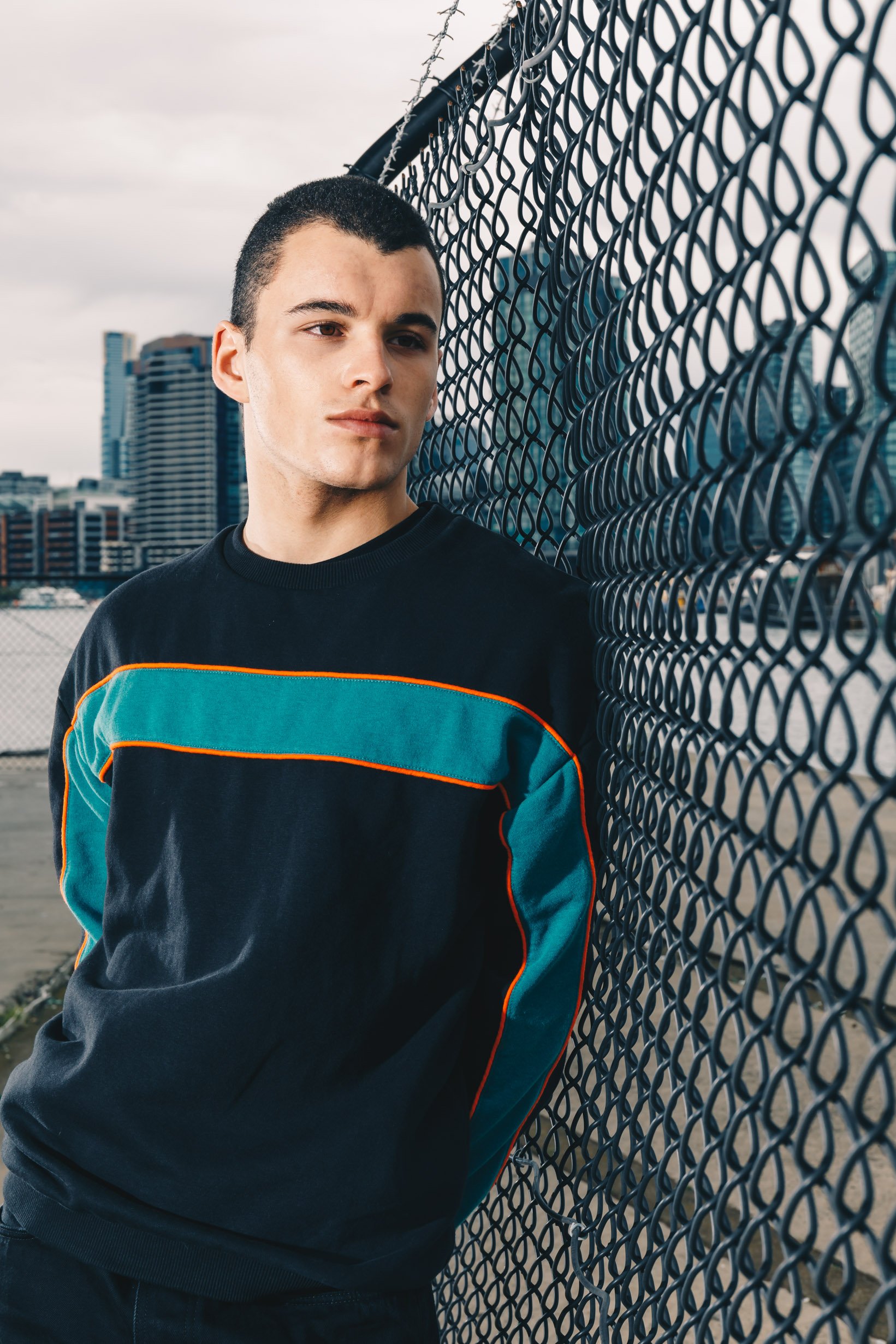 Young man leaning against a chain-link fence with city buildings in the background.