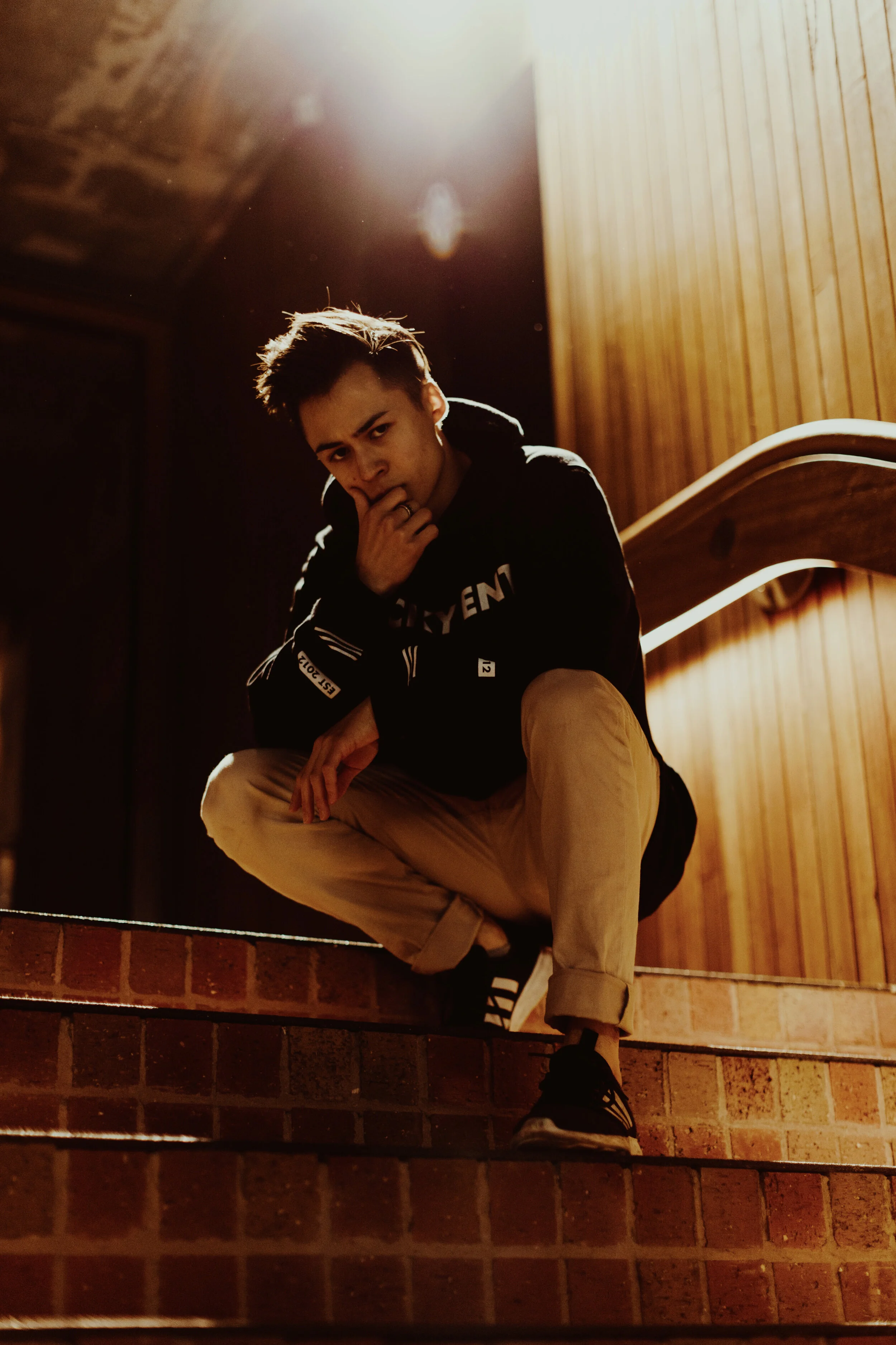 A young man crouching on brick stairs, looking at the camera with hand near his face, in a dimly lit indoor space with wood-paneled wall behind him.