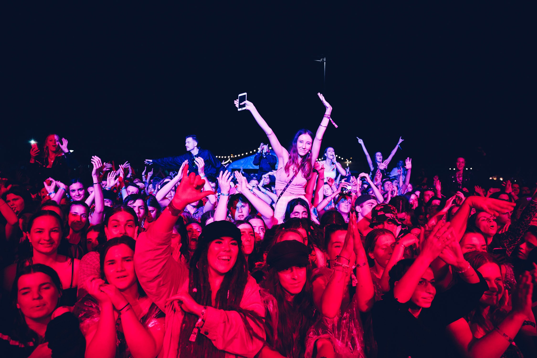 Crowd of people at a music concert or festival at night, with some individuals being lifted above the crowd. The scene is illuminated with colorful lighting, and people are enjoying the live event.