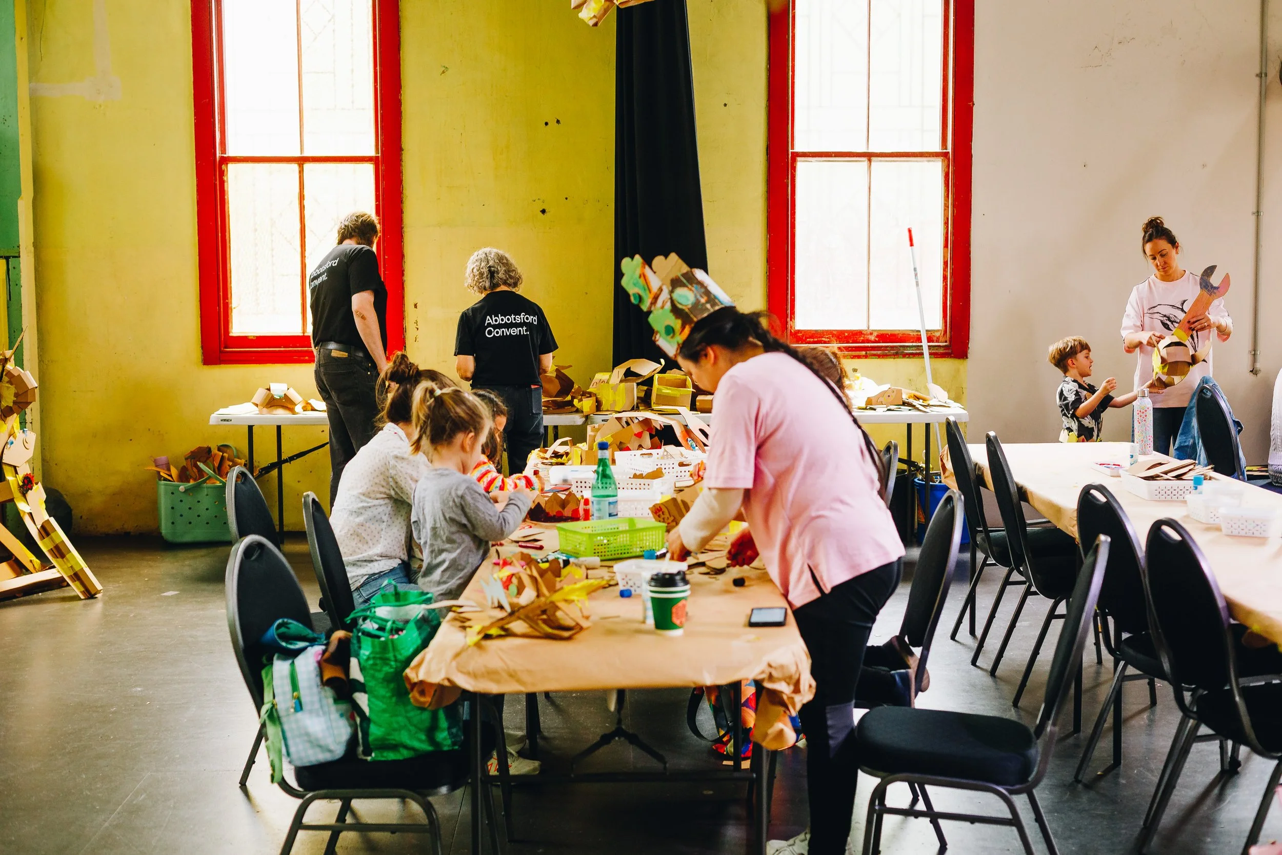Children and adults crafting paper Christmas trees in a bright room with yellow walls and large windows, some wearing shirts with 'Abbotsford Convent' written on the back.