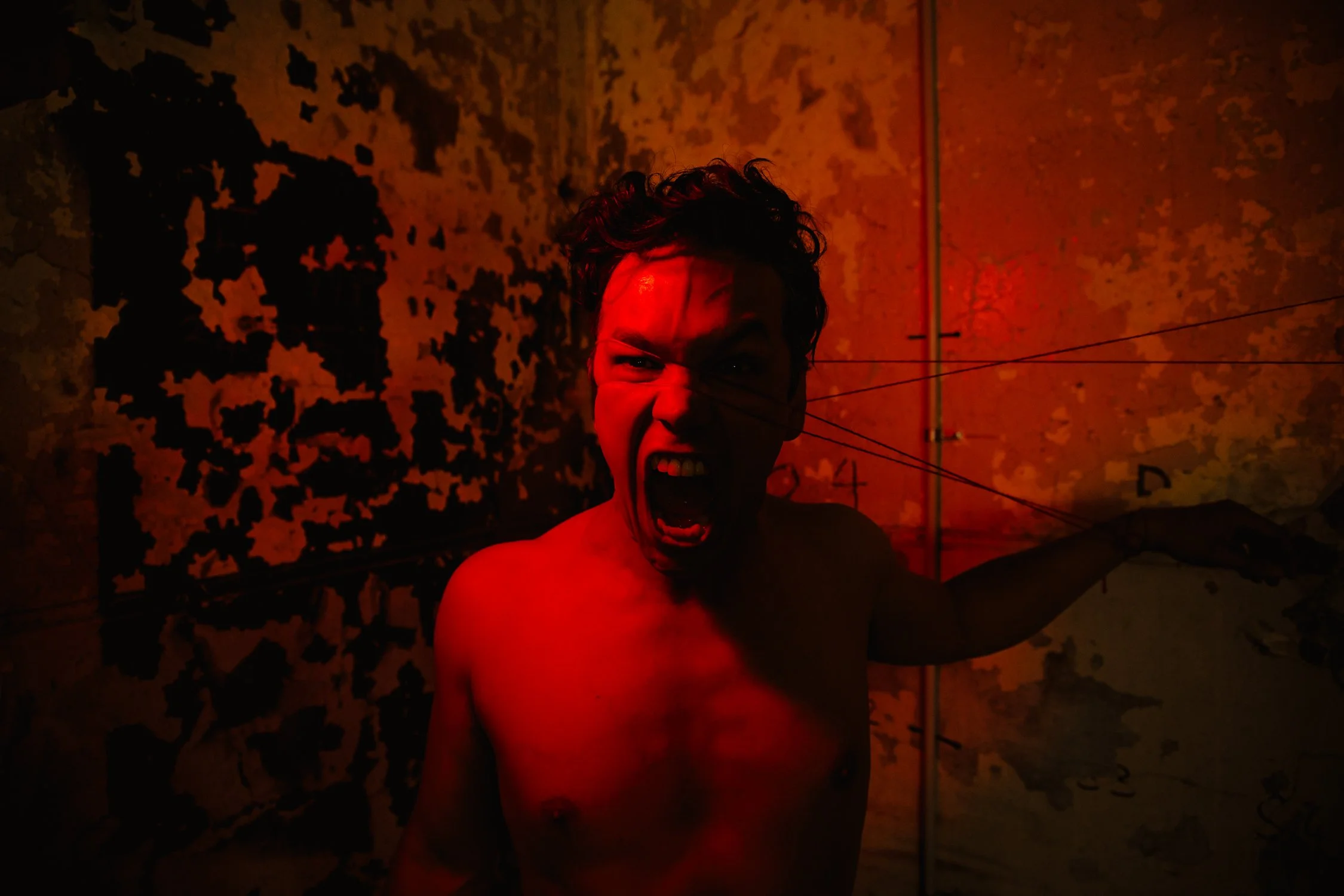 A shirtless man with curly hair screaming in a dark, red-lit room with a peeling wall and technical lines on the wall.