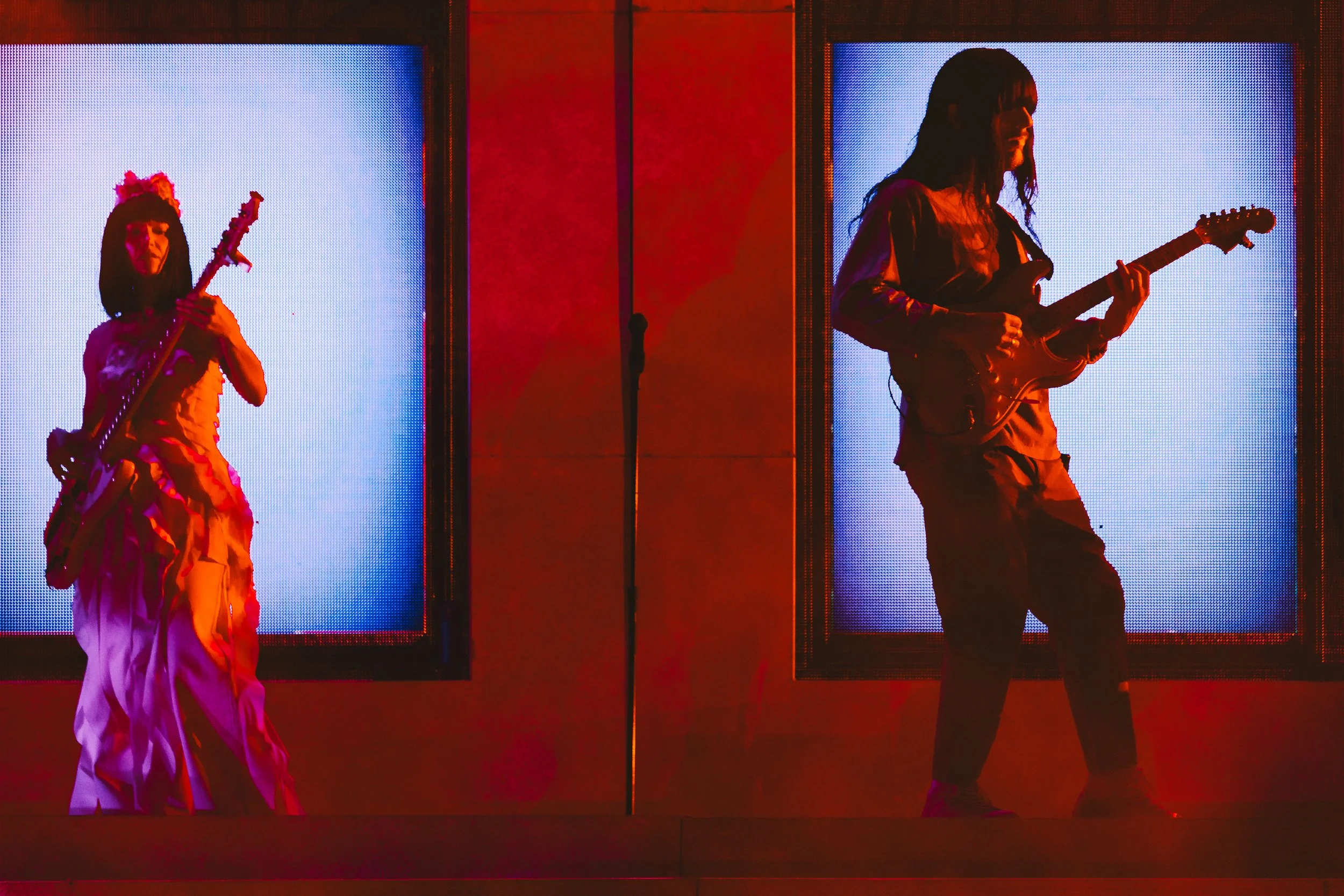 Two women playing electric guitars on a stage with illuminated screens behind them, one with a dress and the other in casual clothing.