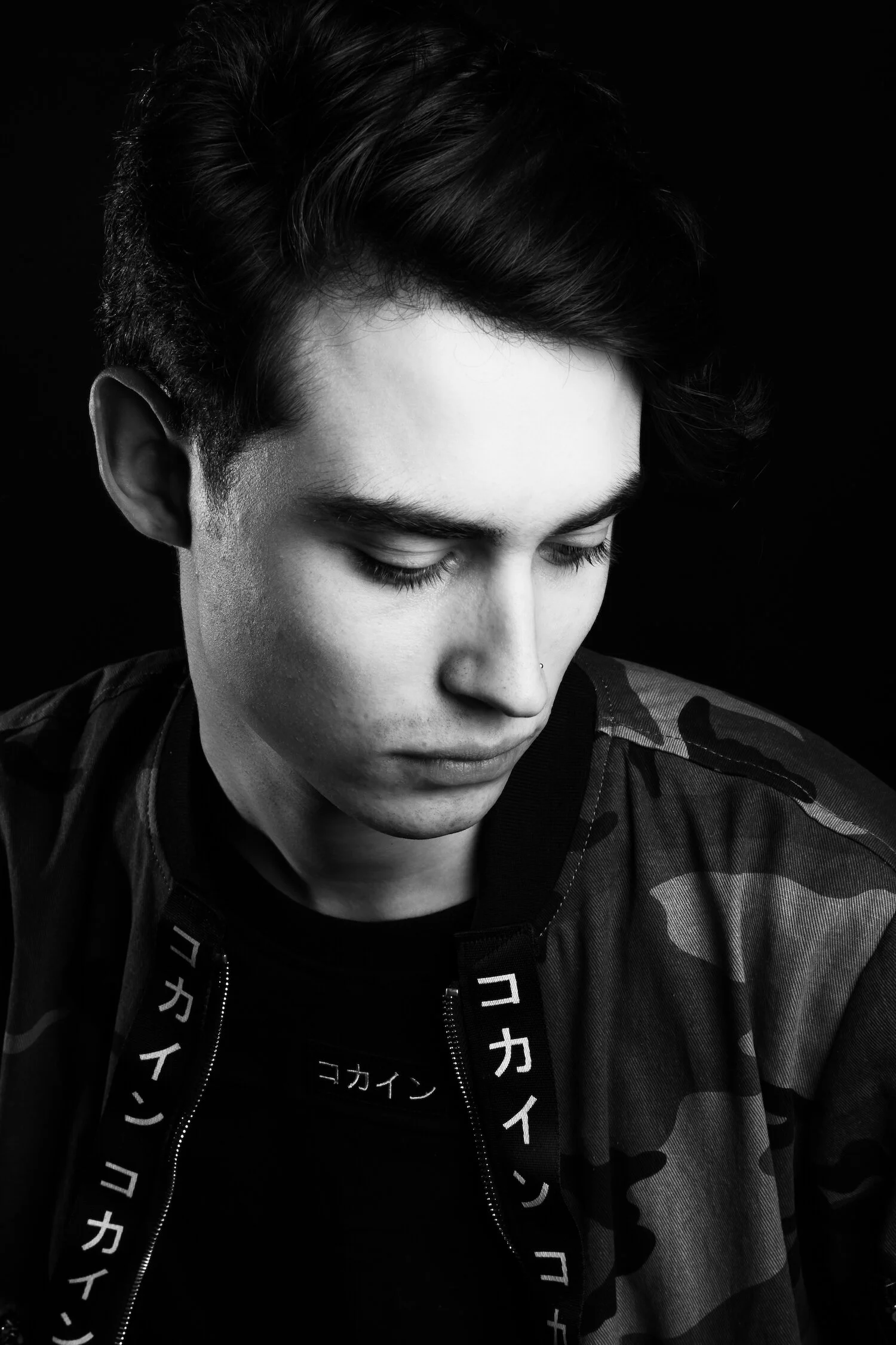 A black and white portrait of a young man with dark hair, looking downward, wearing a camouflage jacket and a shirt with Japanese characters.