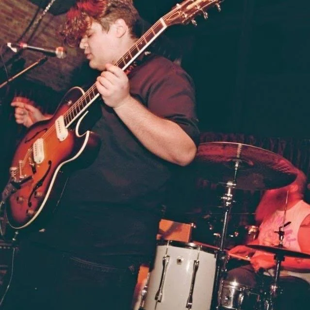 A young man playing an electric guitar during a live music performance, with a drummer visible in the background.
