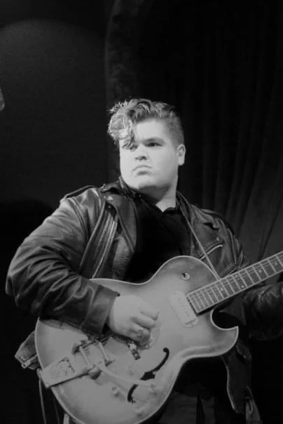 Young man with styled hair wearing a leather jacket, playing an electric guitar on stage with a dark curtain backdrop.