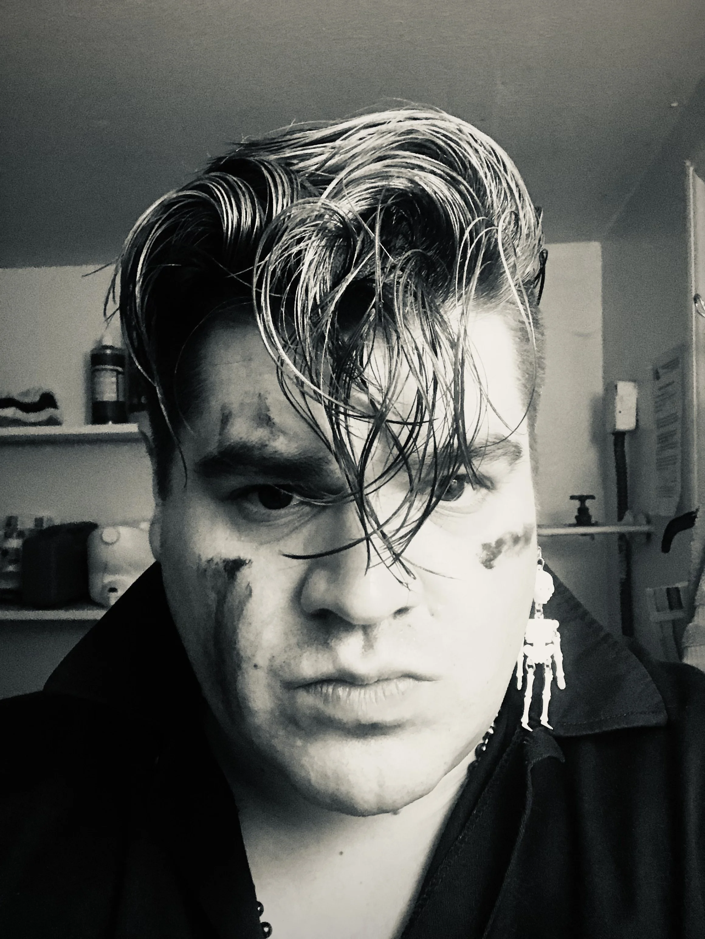 A black and white portrait of a person with wet, styled hair, wearing earrings, and a dark shirt. The background shows a room with shelves and various objects.