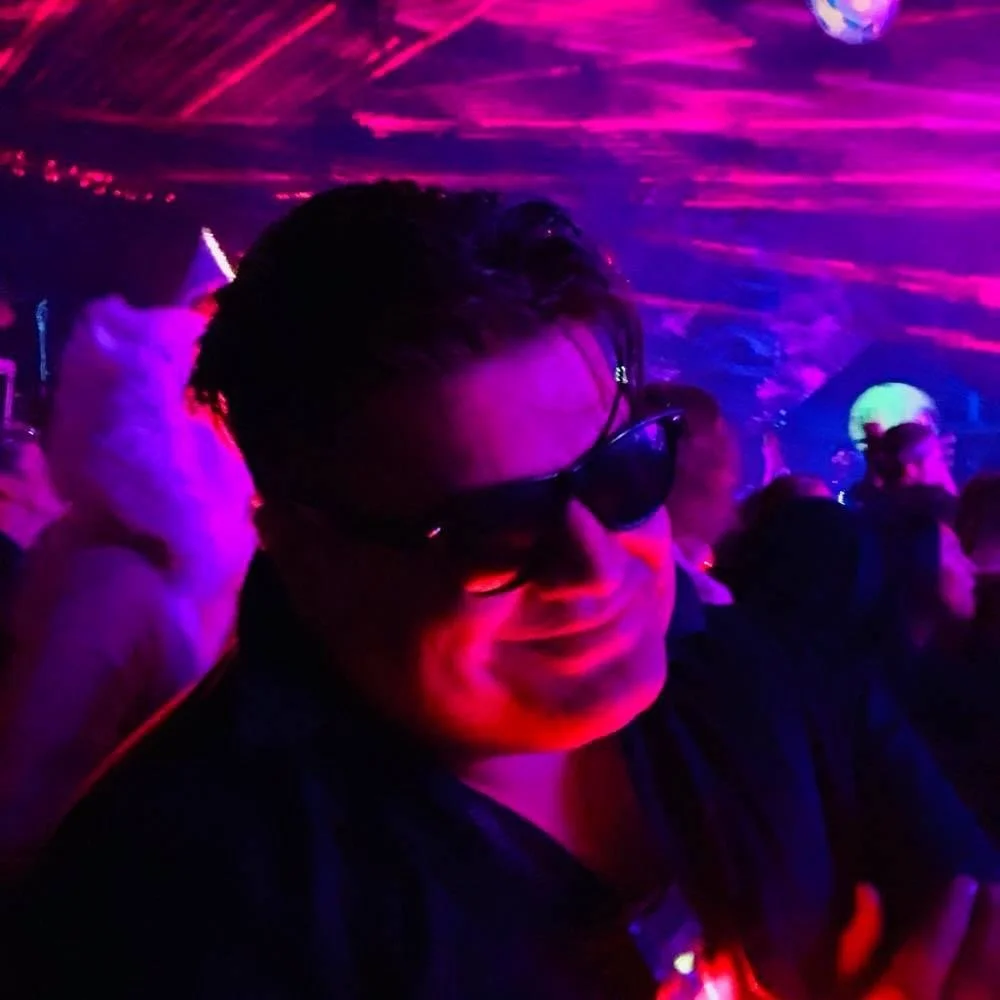 A smiling person wearing sunglasses in a dark, colorful party scene with vibrant pink, purple, and blue lights and other people in the background.