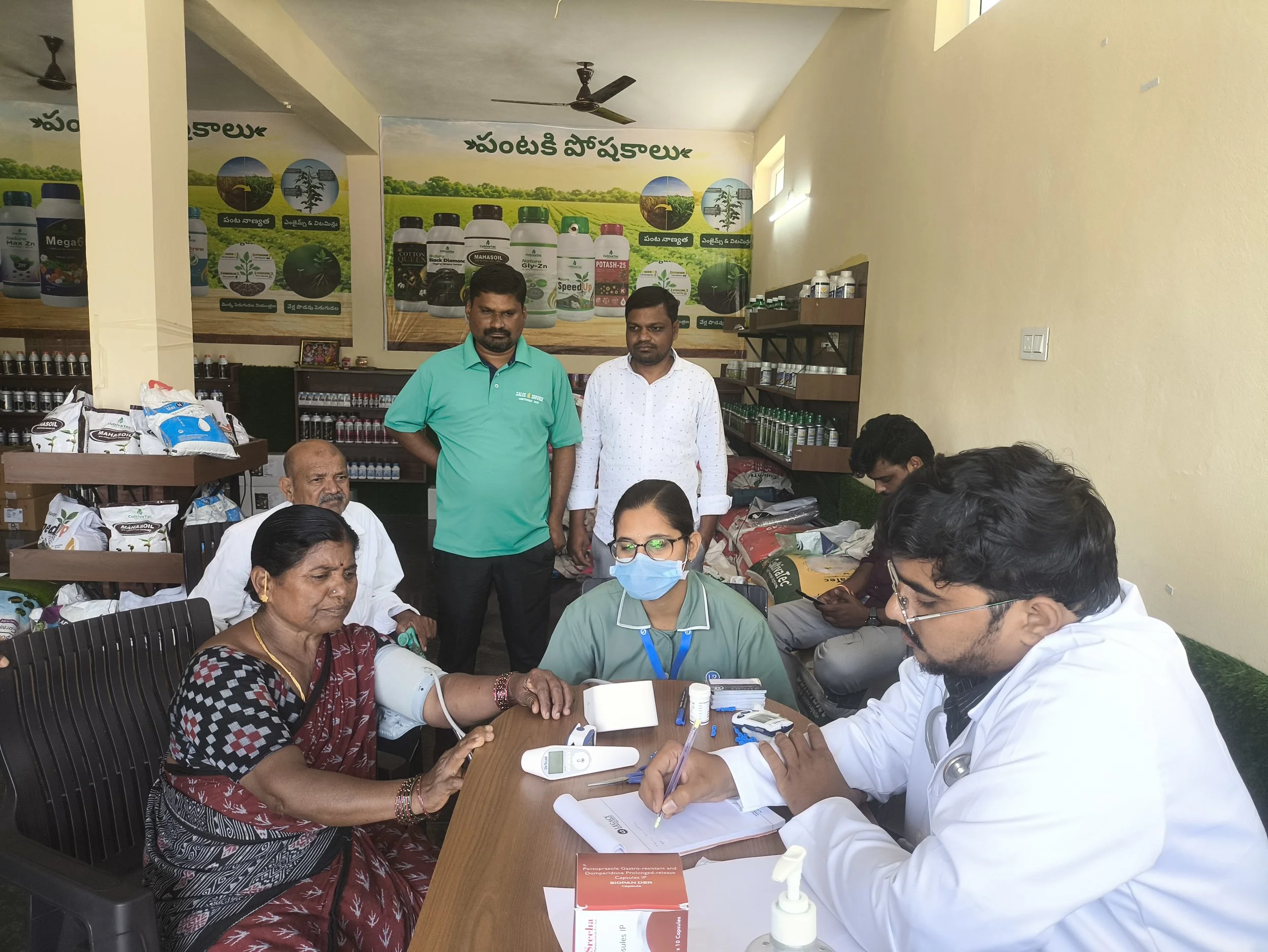 A medical consultation taking place in a room with various agricultural product posters on the wall. An elderly woman is getting her blood pressure checked by a healthcare worker, while other people and healthcare professionals are present.