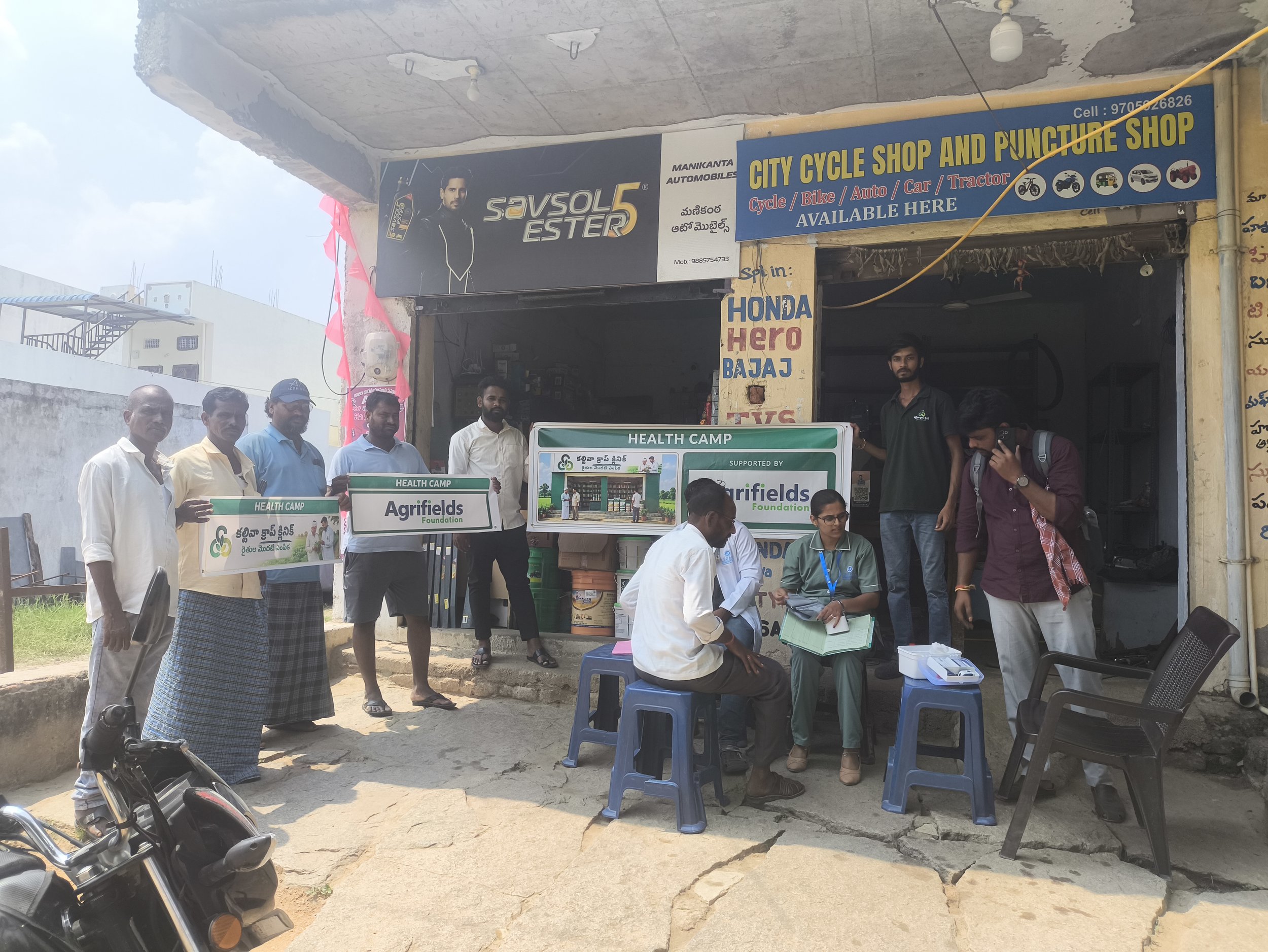 Group of people standing and sitting outside a shop, holding signs for Agrifields Foundation. The shop in the background has signs for a city cycle shop and Puncture shop, with various vehicles depicted and contact details. The shop has a large poste