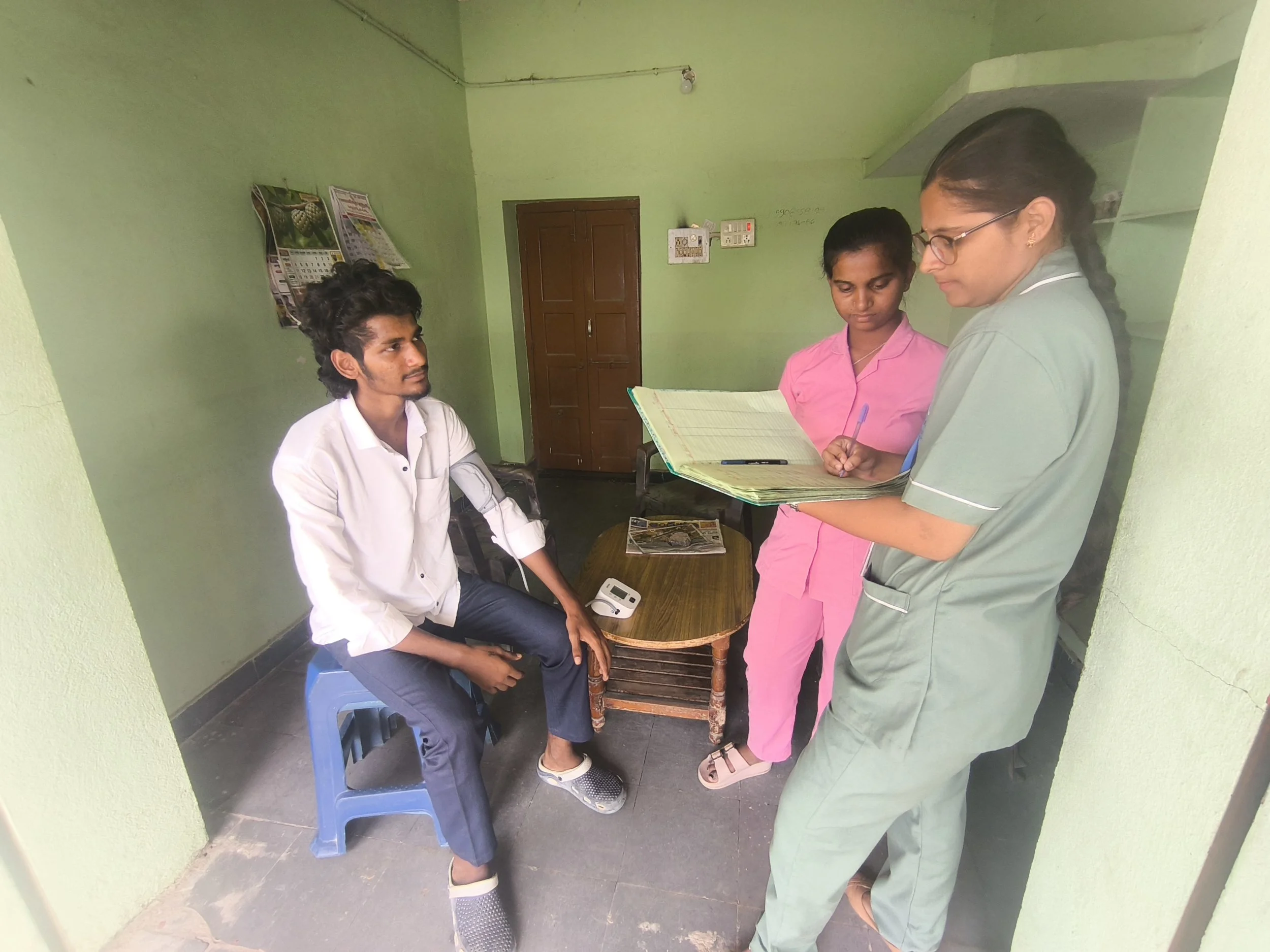 A man sitting on a stool in a clinic has his arm checked by a nurse with glasses. Two other women are standing next to him, one holding a notebook and pen, and the other looking on. The room has green walls and a wooden door.