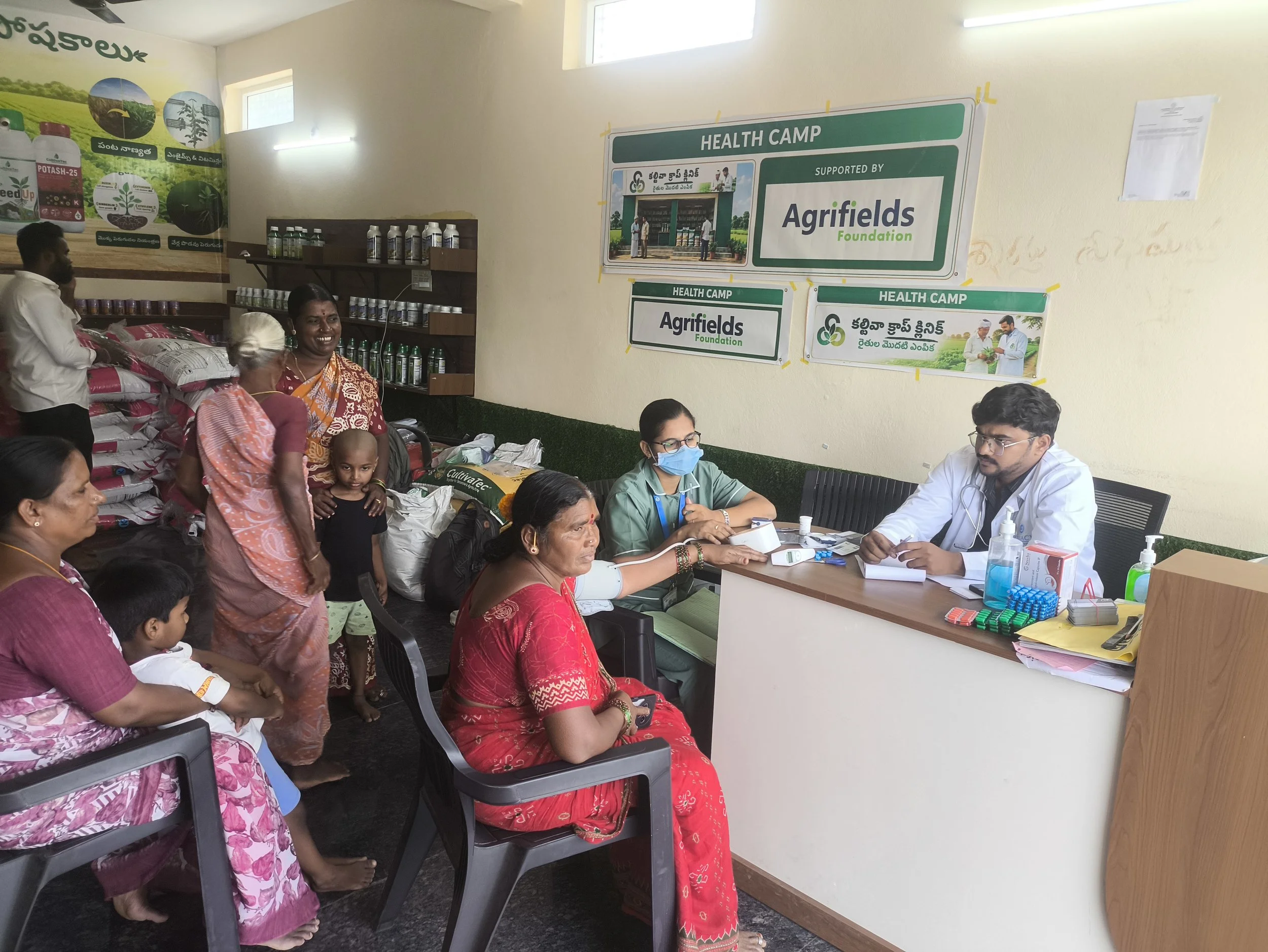 Farmers gathered at an Agrifields Foundation health camp, with a healthcare worker wearing a blue face mask seated at a table with medical supplies.