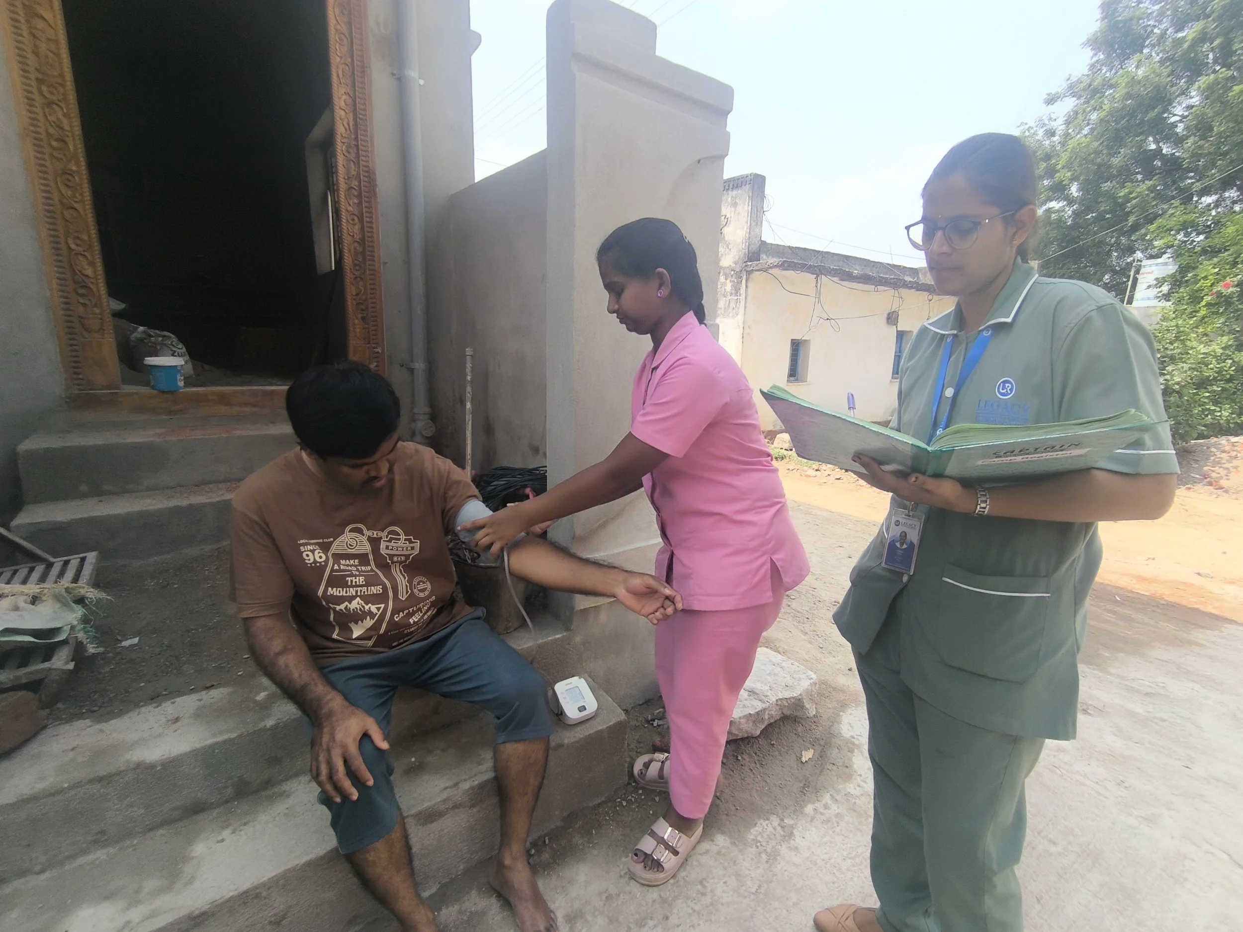 A woman in pink scrubs and a man with glasses and a uniform are attending to a young man sitting on the steps outside a building. The young man appears to be receiving medical attention.
