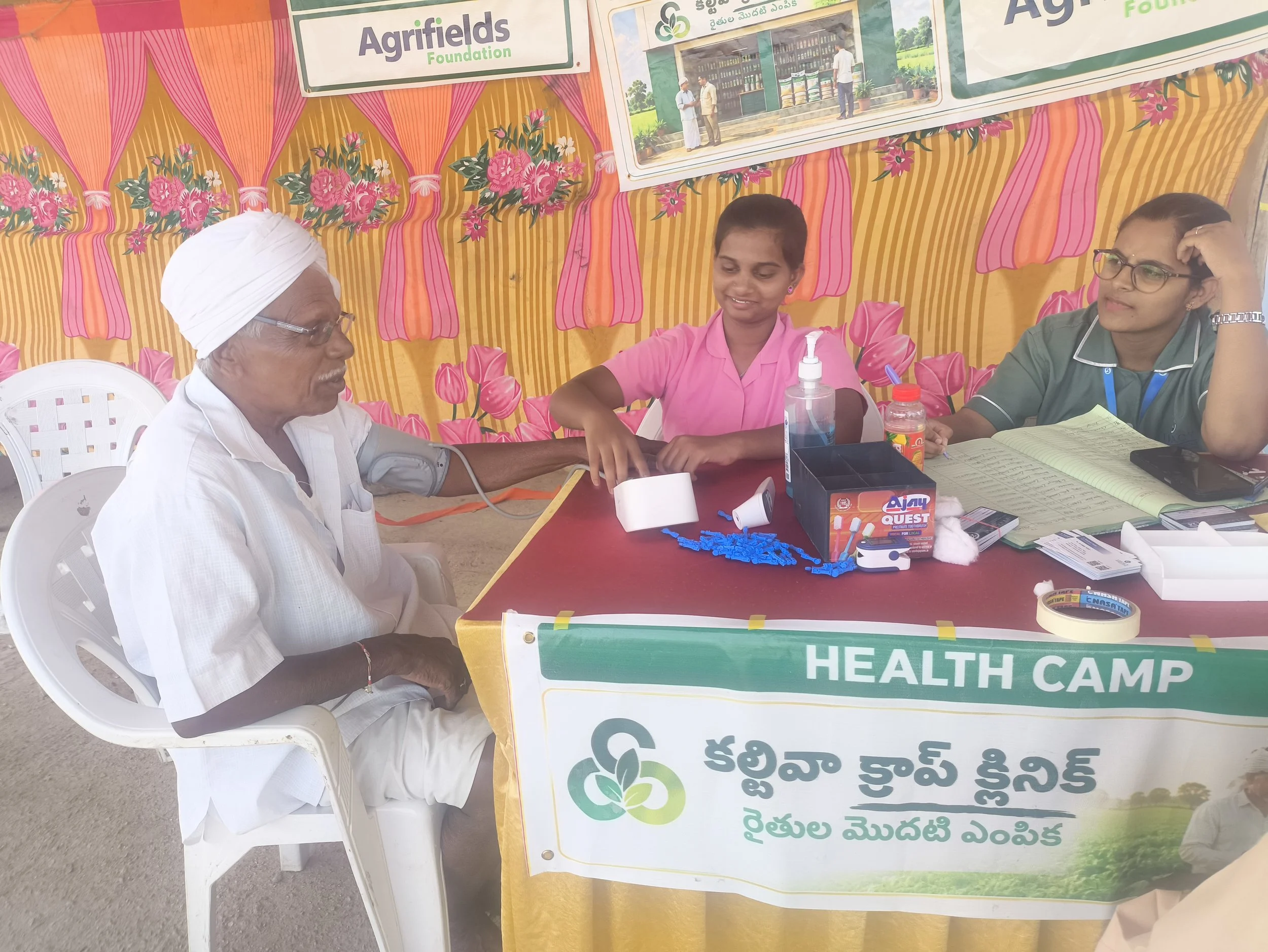 Three women and an elderly man seated at a table during a health camp, with colorful floral and striped fabric draped behind them.