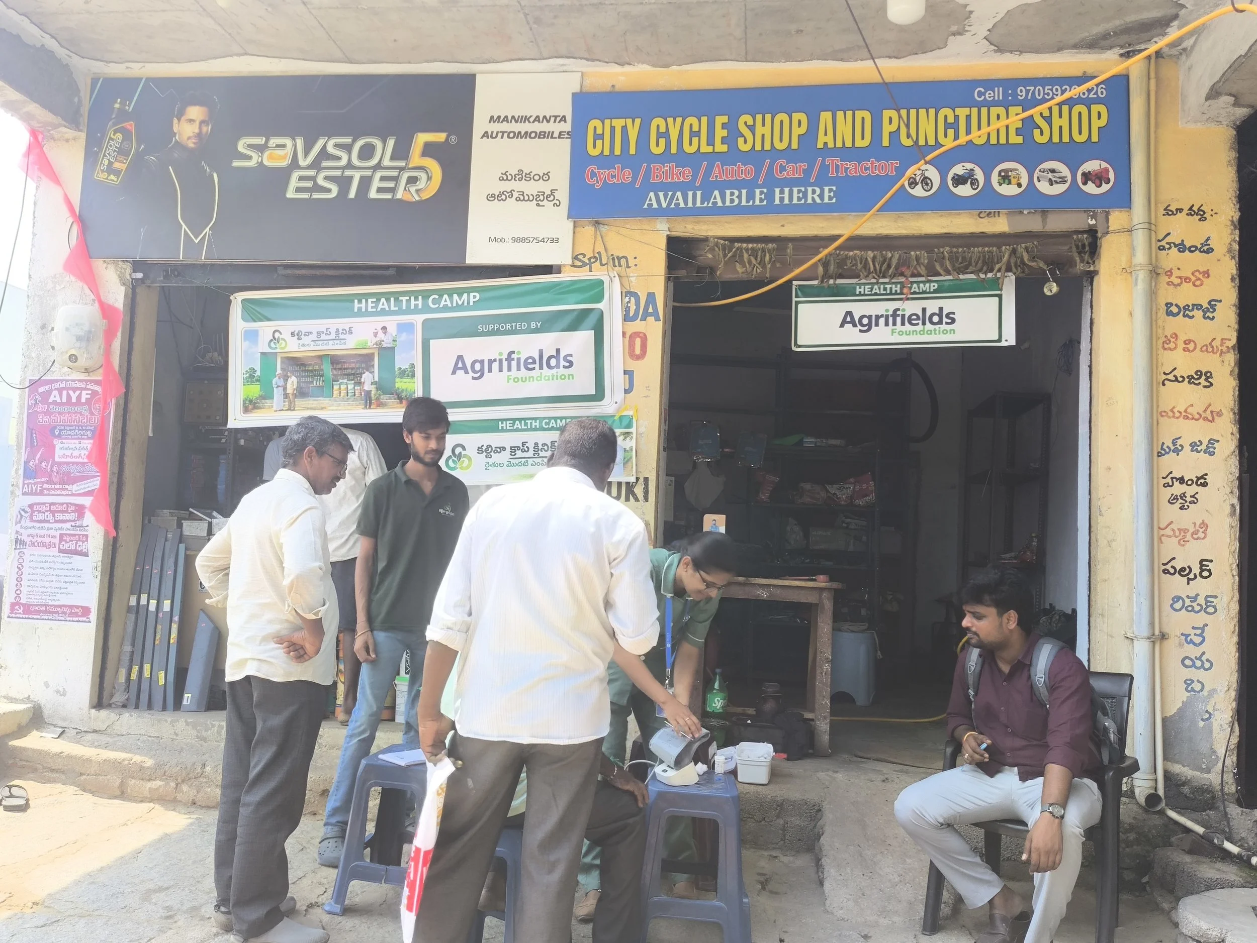 People gathered outside a shop with signs indicating it sells auto parts, motorcycles, bicycles, and offers health camp services, with some individuals sitting and others standing.