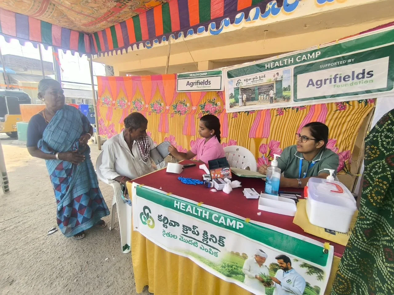 A health camp setup at an outdoor event with a colorful striped canopy. Several people are seated at tables with medical staff checking a man's blood pressure. There are banners with the text 'Health Camp' and 'Agrifields Foundation.' Medical supplie