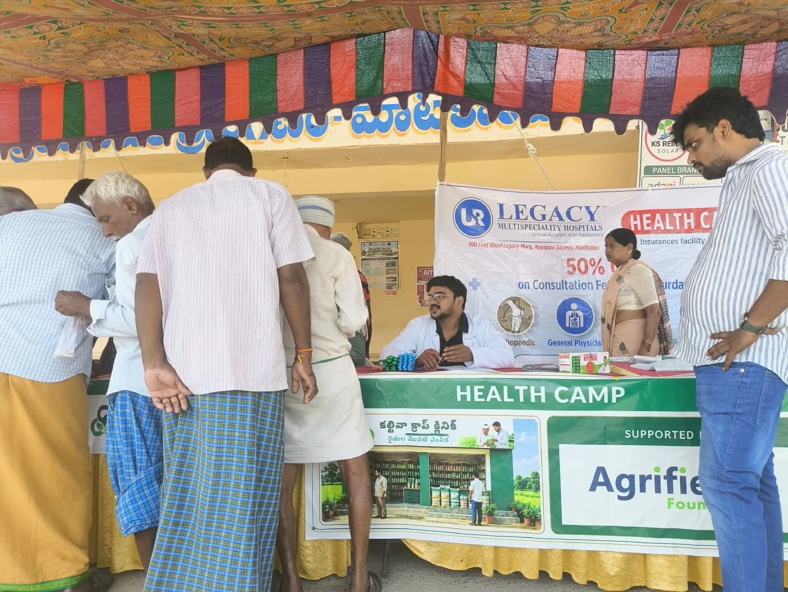 A health camp booth at an outdoor event with several people gathered around. The booth has a banner advertising health check-ups and consultation discounts, with one person sitting at the table managing registrations or inquiries and others standing 