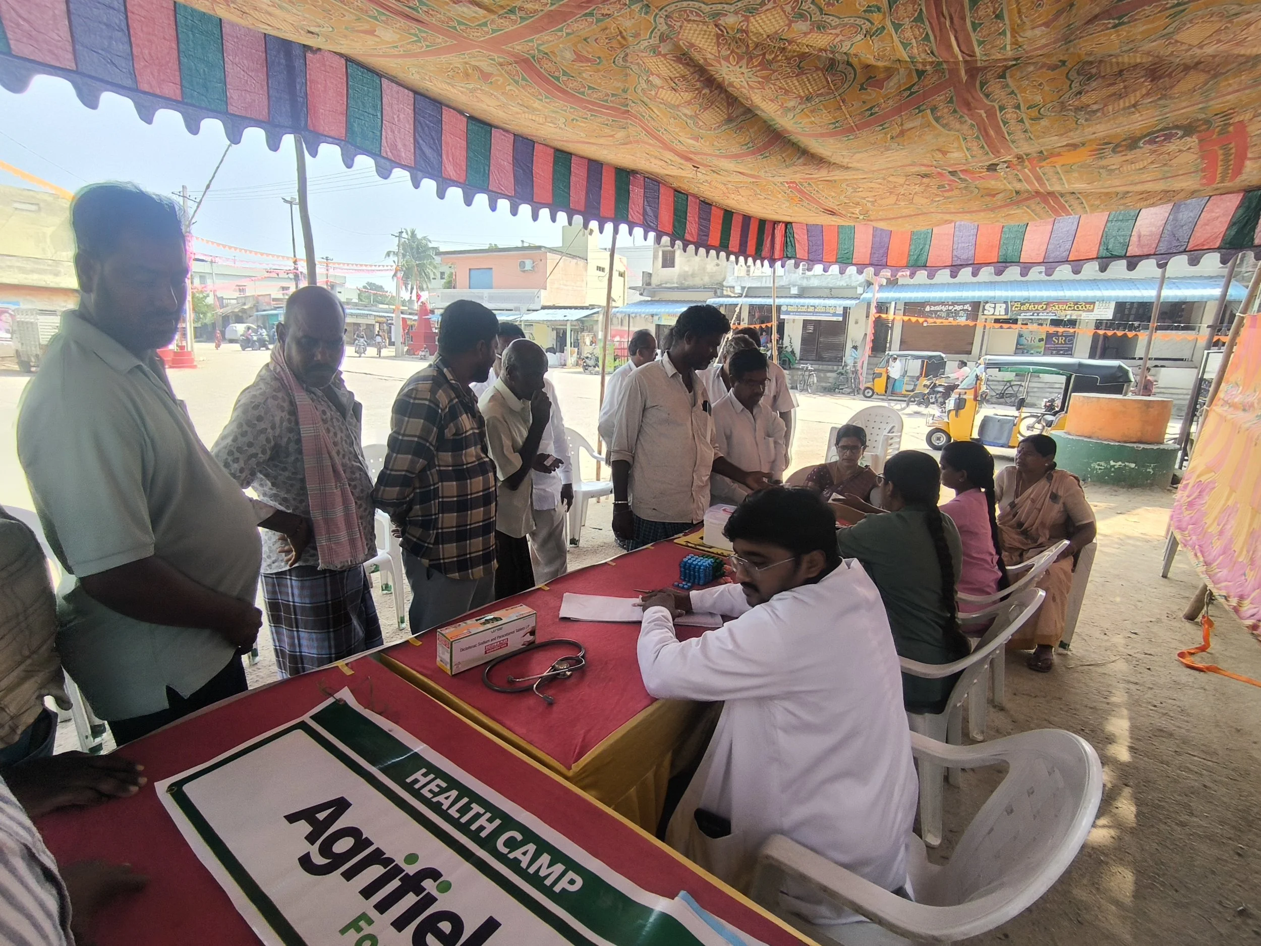 A group of people gathered around a table at an outdoor health camp during daytime. The table has health-related materials, including a box labeled 'Health Camps'. Several men are standing and sitting, engaging in conversation, with background of sho