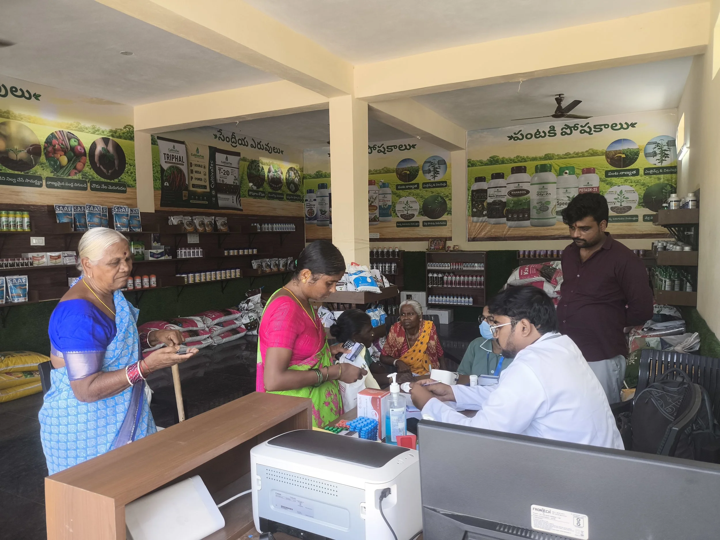 Farmers gathered around a table inside an agricultural supply store, with shelves of farming products and Telugu posters featuring crops in the background.