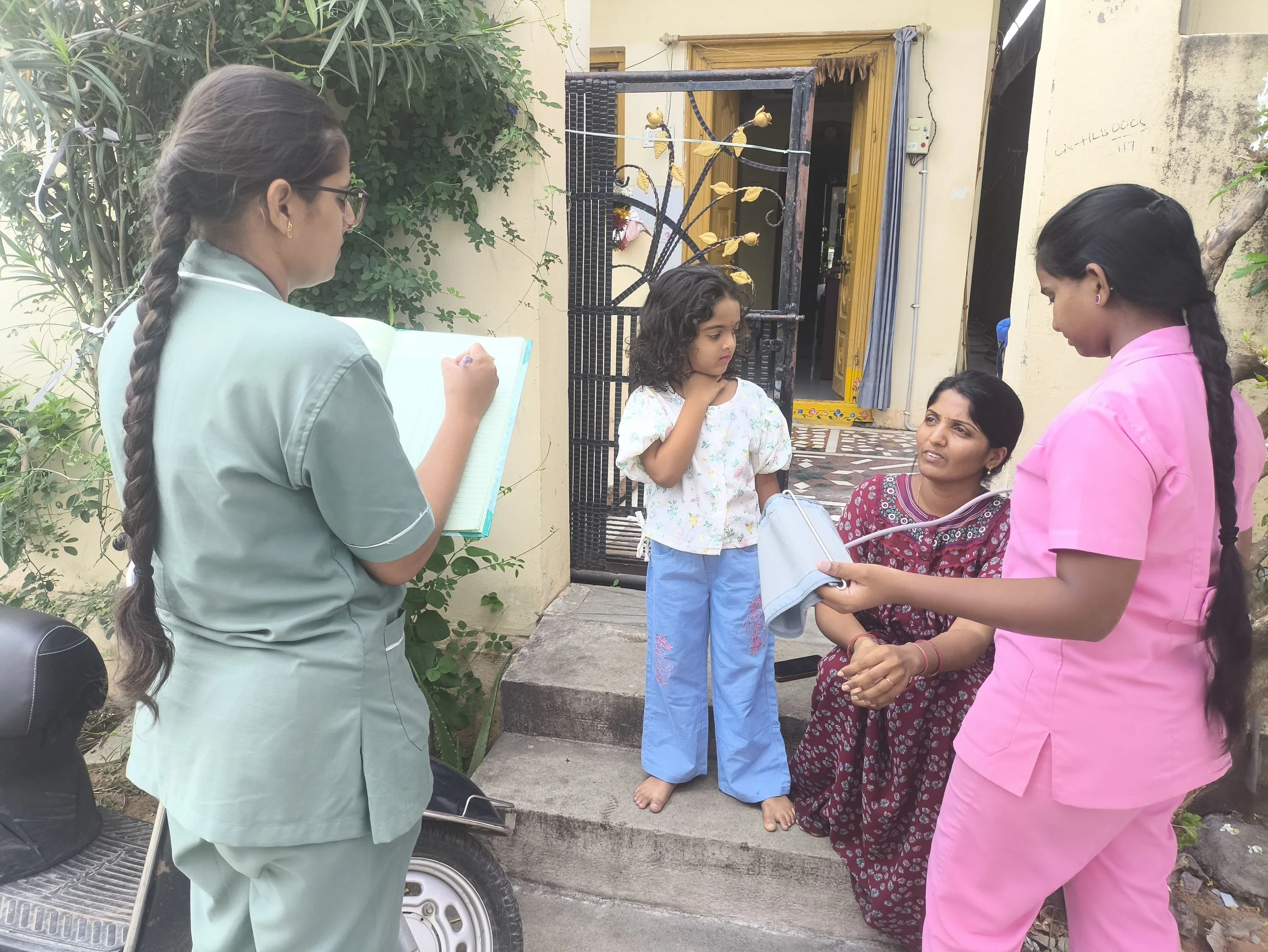 A group of women and a young girl are gathered outdoors near a house entrance. One woman is holding a notepad and pen, another is taking notes, and the young girl appears to be explaining something. The group seems engaged in a discussion.
