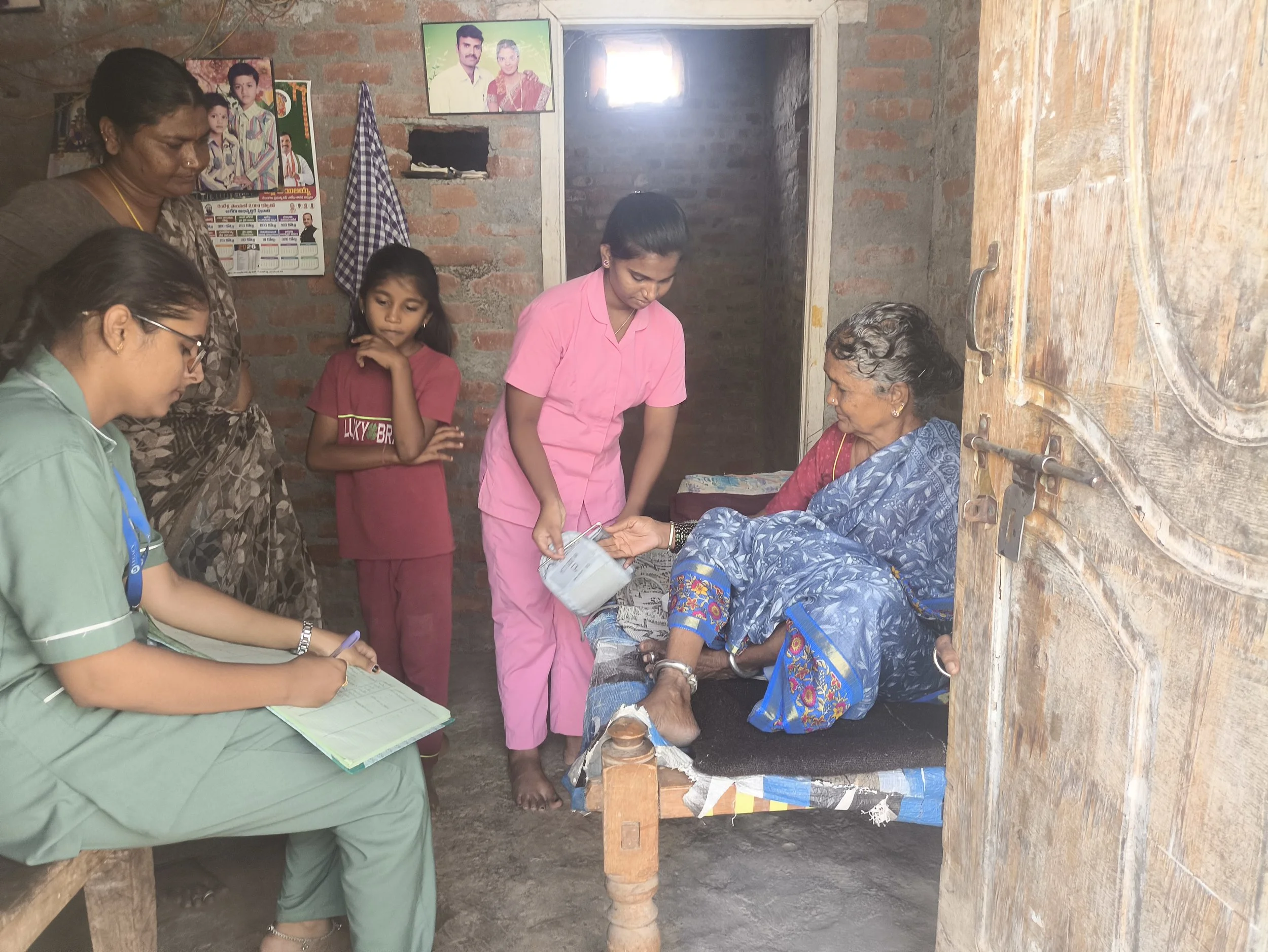 Group of women and a young girl inside a simple brick-walled room, with one woman sitting on a wooden bench, others standing, and a woman in pink serving water from a jug; pictures on the wall and a wooden door partially open.