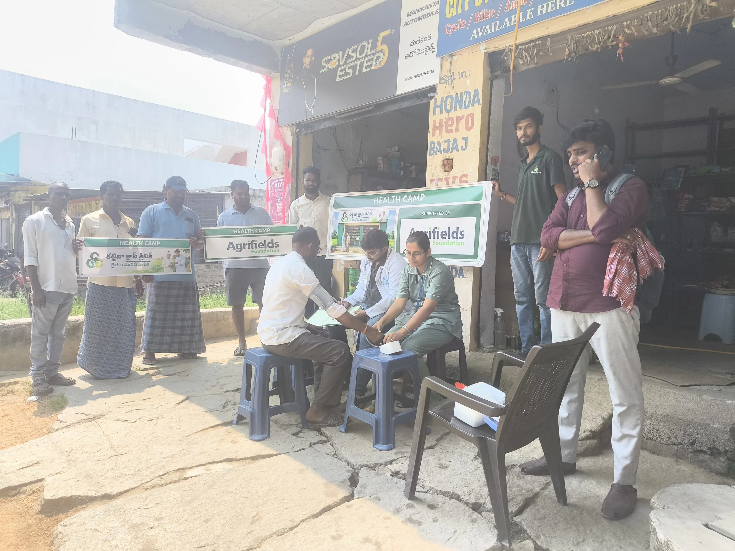 People gather outside a shop, some holding banners that say 'Health Camp' and 'Agrifields Foundation.' A healthcare worker examines a man, with several others standing nearby. The shop has signs for various products and services, with some individual