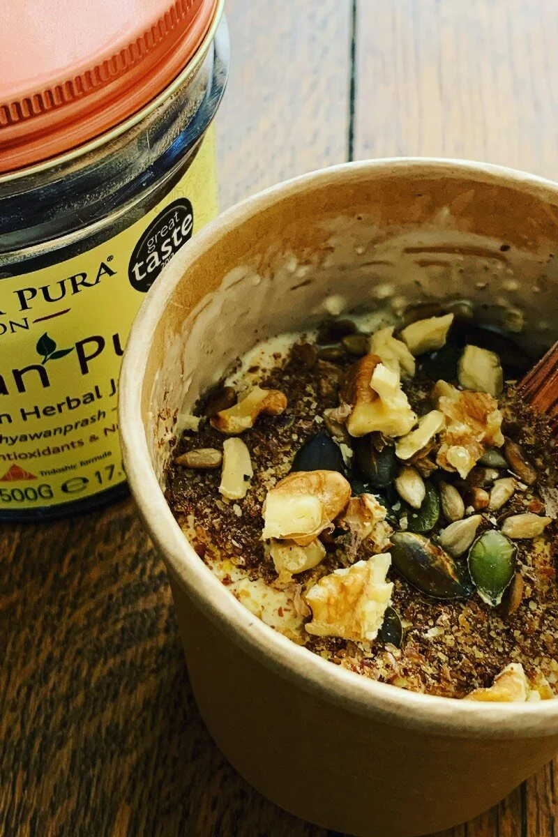 A cup of yogurt topped with mixed nuts and seeds, with a jar of herbal supplement powder nearby, on a wooden surface.