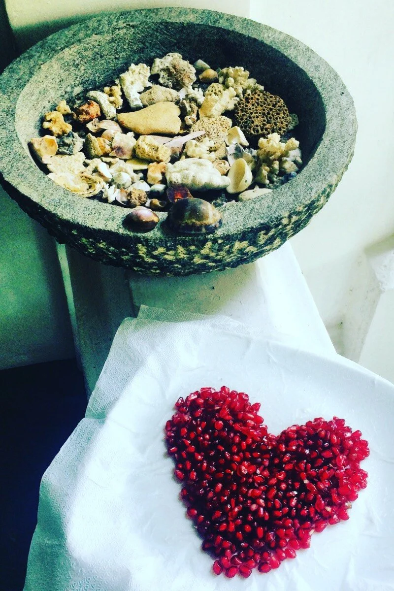 A stone bowl filled with various seashells and coral pieces, and a separate white napkin with pomegranate seeds arranged in the shape of a heart.