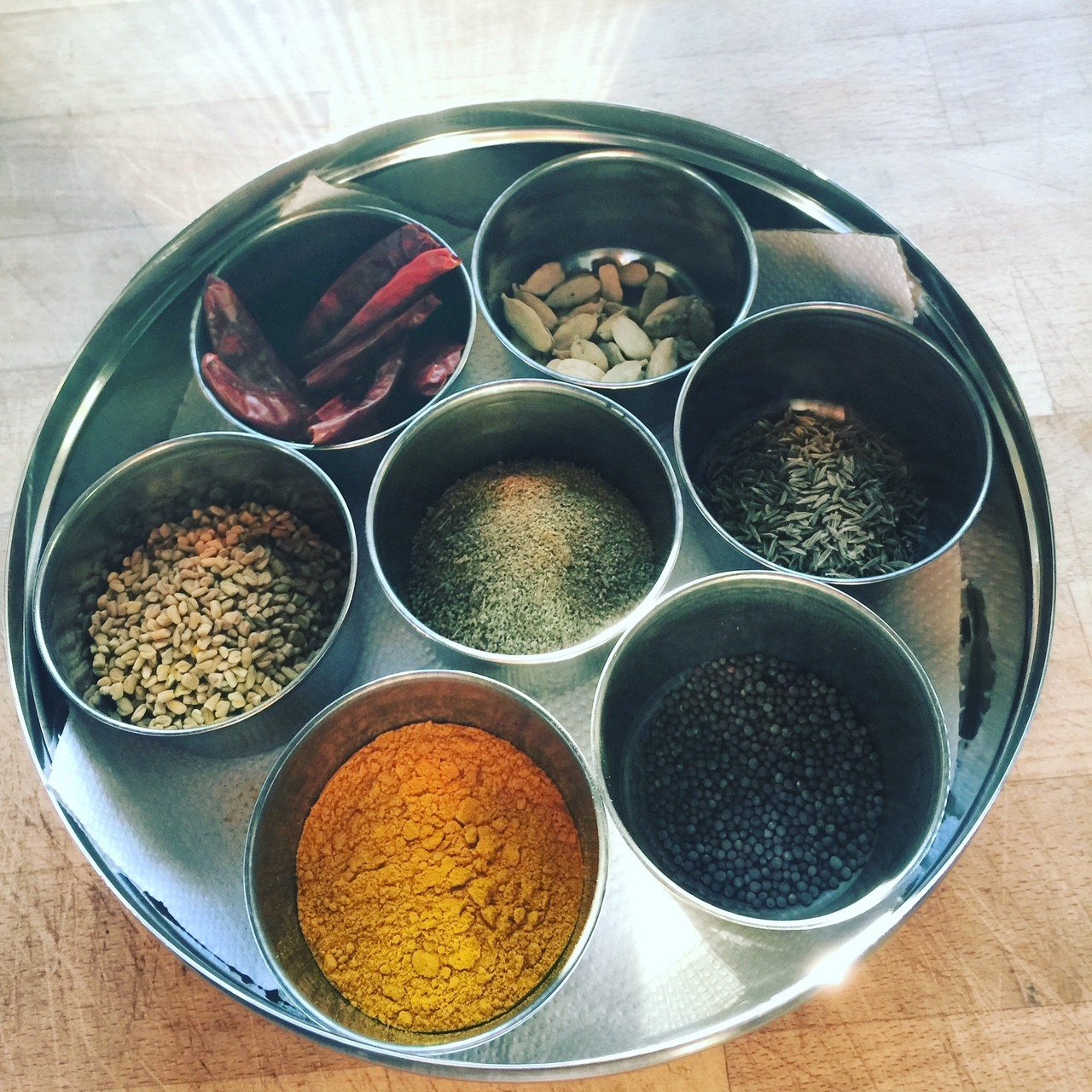 Metal tray with various spices and herbs in small containers, including dried chili, pistachios, sunflower seeds, cumin, turmeric, and black mustard seeds.