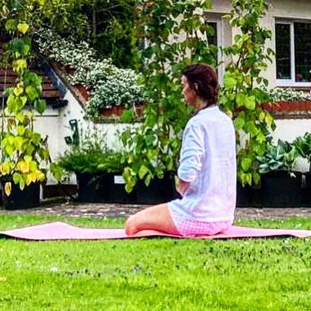 A woman in casual clothing practicing yoga or meditation on a pink mat outdoors in a garden.