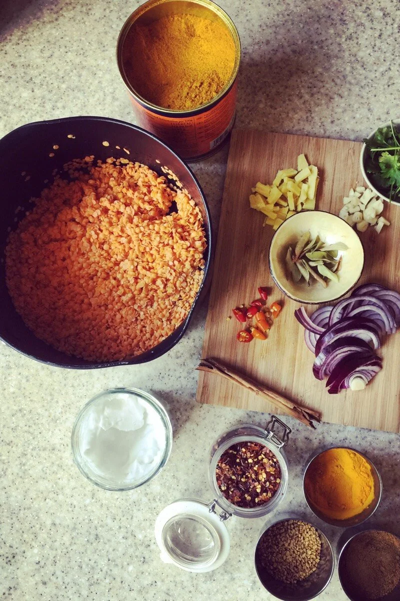 Uncooked red lentils in a bowl, with various spices and chopped vegetables on a cutting board, including onion, ginger, garlic, red chili, and cilantro, along with cooking ingredients like turmeric powder, ground spices, lentils, sour cream, and oil,