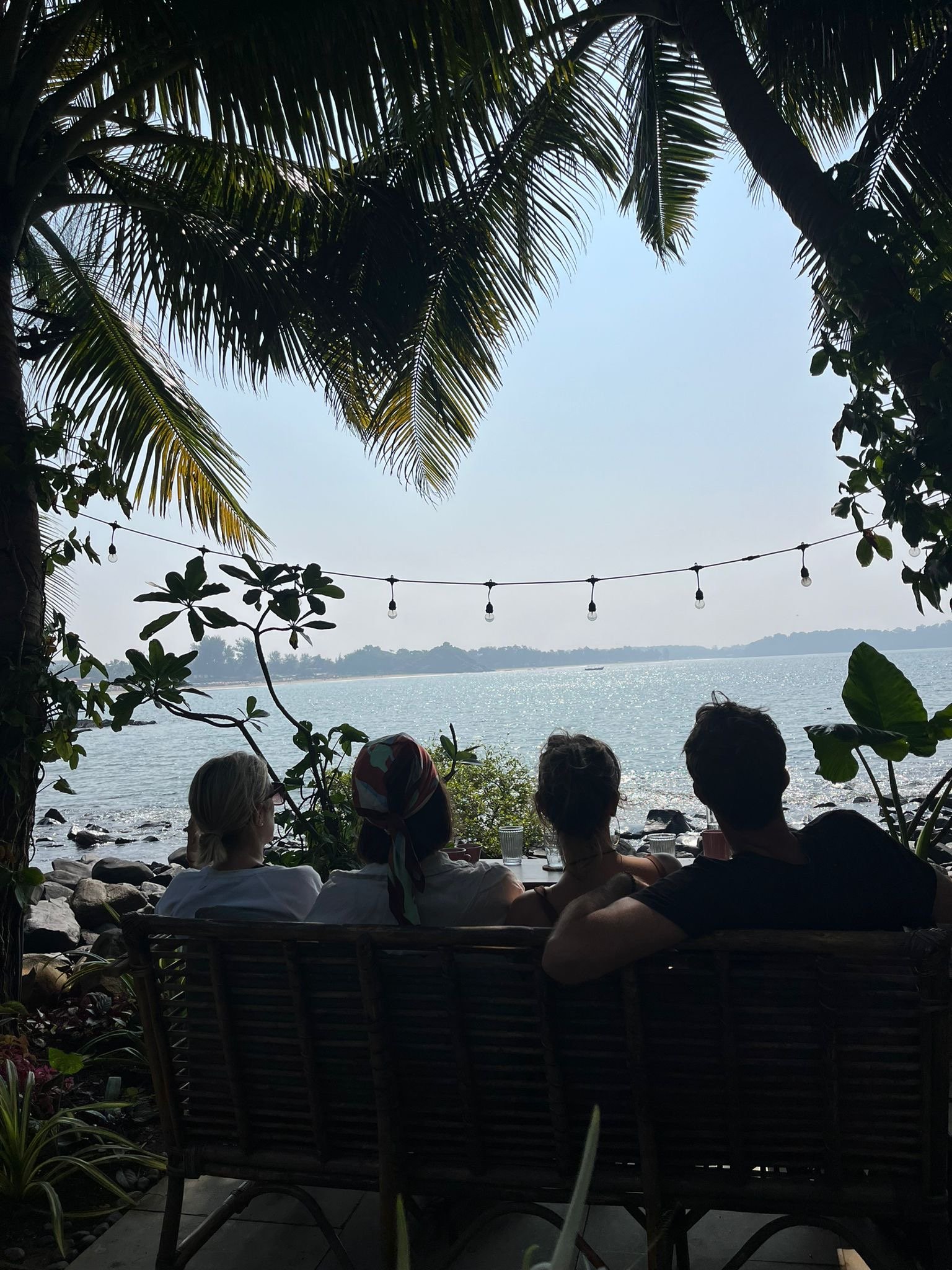 Four people sitting on a bench under palm trees in Goa India, overlooking a calm body of water with string lights hanging above.