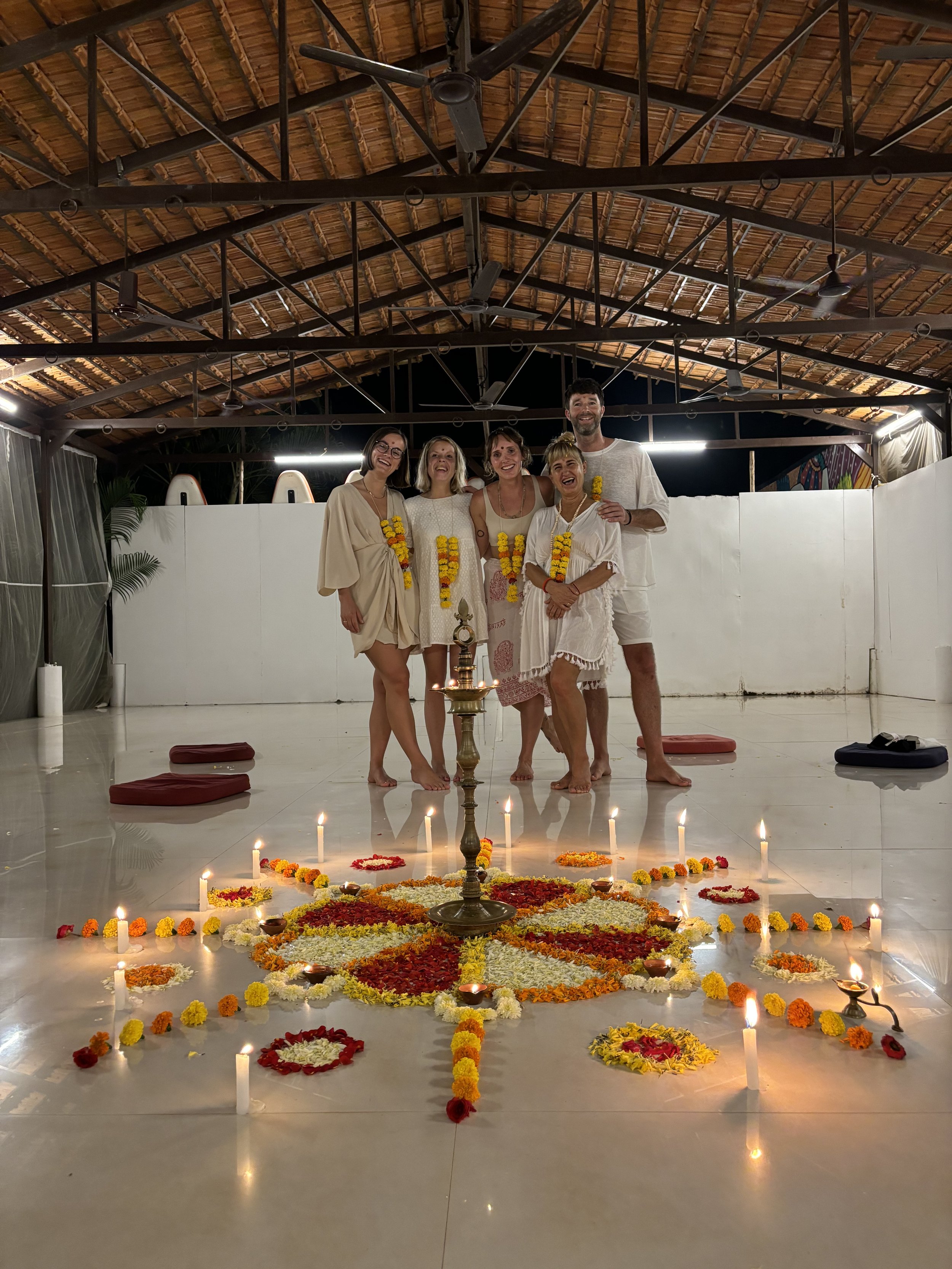 Our Yoga retreat group in Goa India standing in an open-air pavilion with a flower and candle arrangement on the floor. They are dressed in light-colored clothing and wearing garlands.
