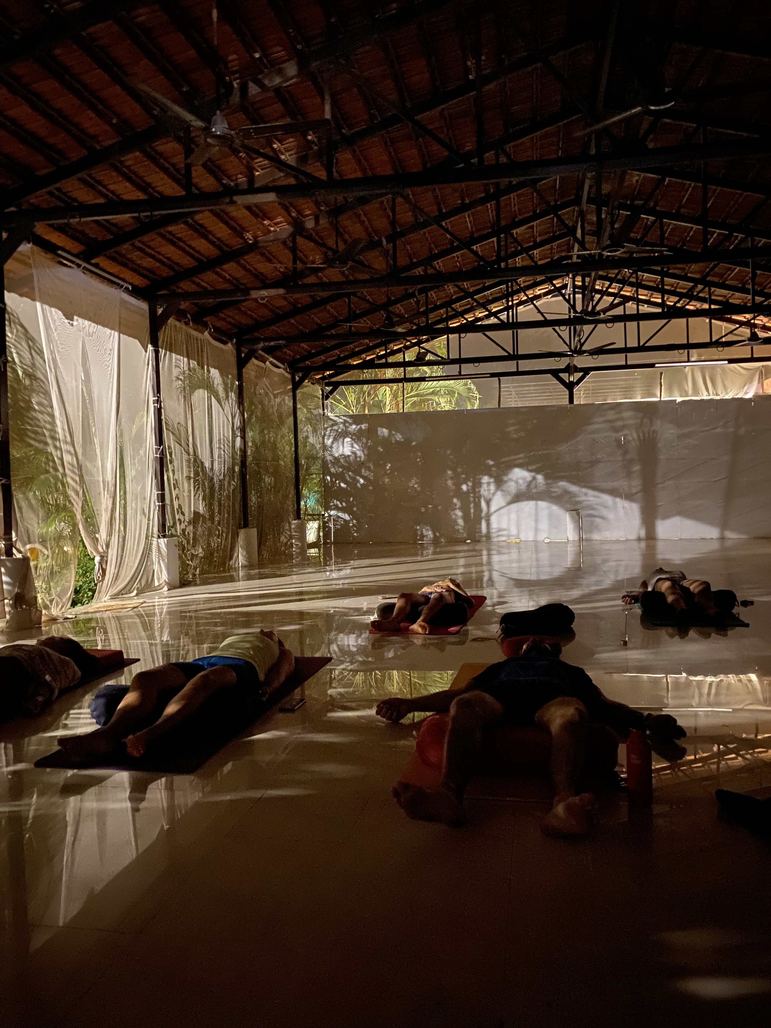 Yoga class in a dimly lit, large open space with high wooden ceilings. Participants are lying on yoga mats in a relaxed pose, likely practicing Shavasana. Sunlight filters through sheer curtains, casting shadows on the floor.