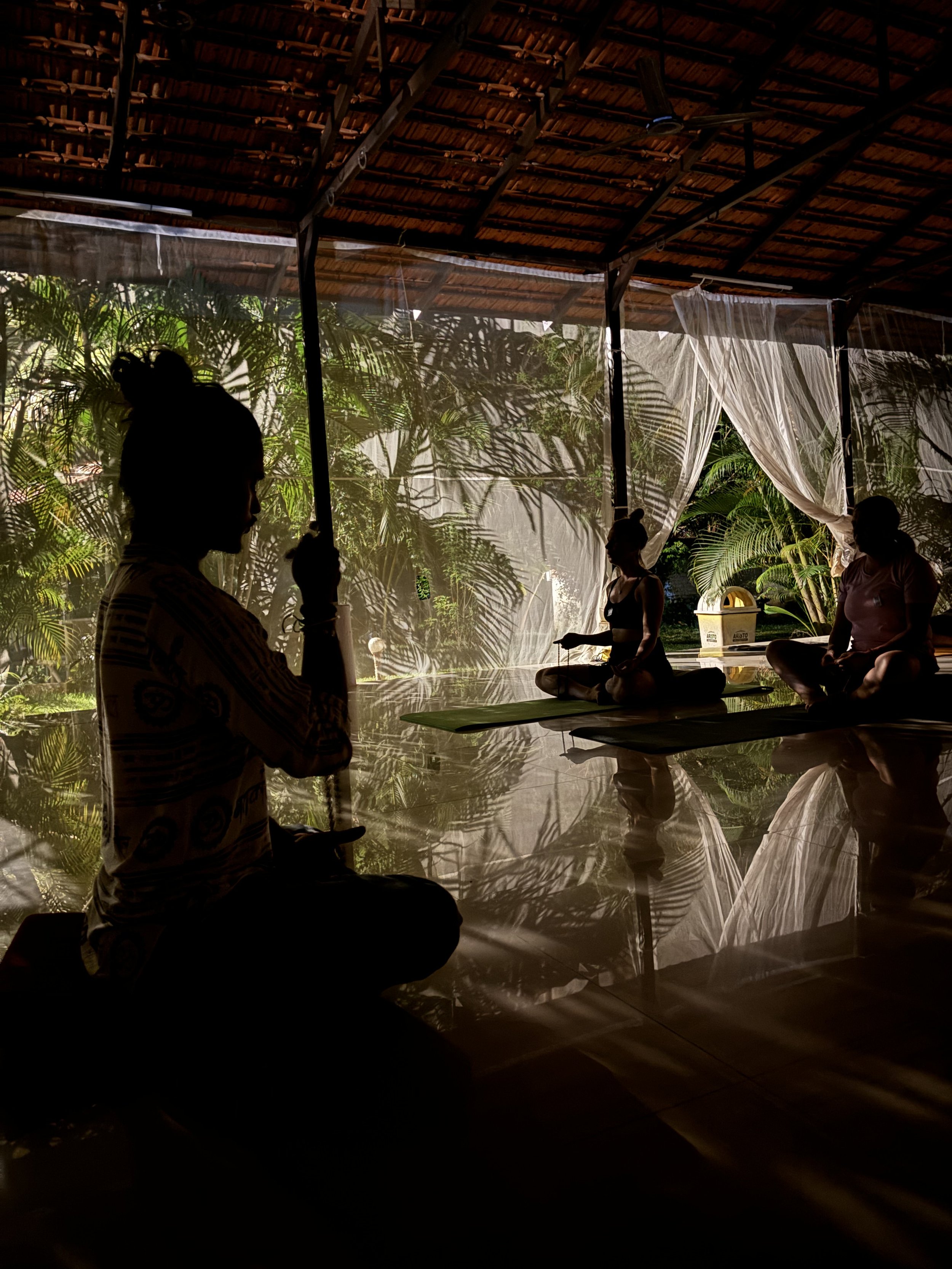 Silhouettes of three people meditating in our Yoga retreat in India Goa in a room with bamboo roof and curtains, surrounded by tropical plants, in a meditative or yoga pose in Goa India