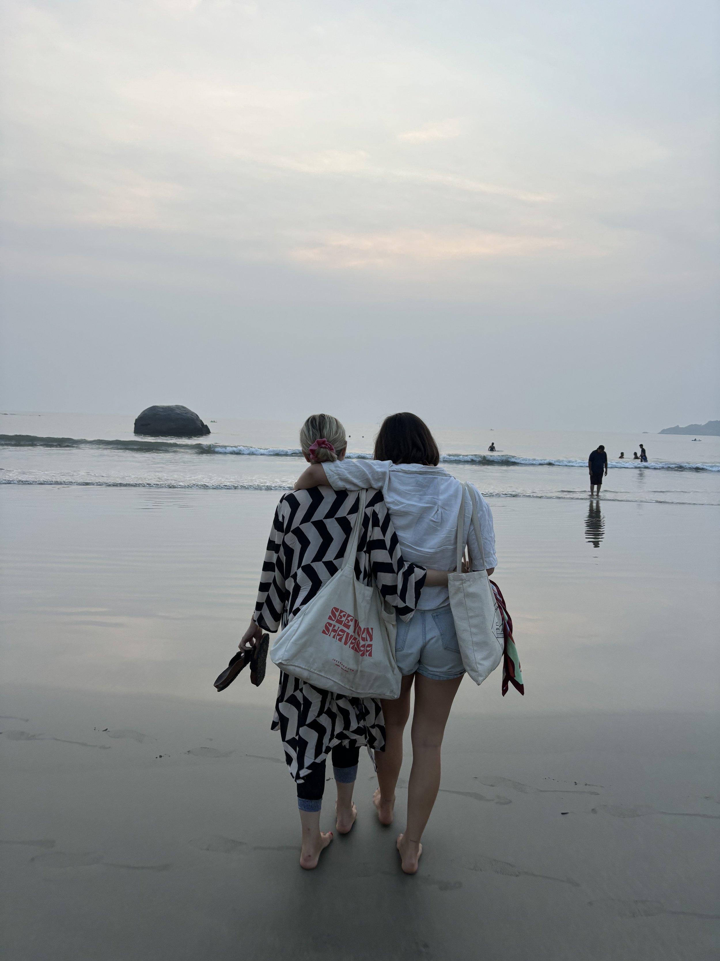 Two people walking on a beach at sunset at Palolem in south goa India, with their arms around each other and carrying bags. They are barefoot, and the ocean and a rocky outcrop are visible in the background.