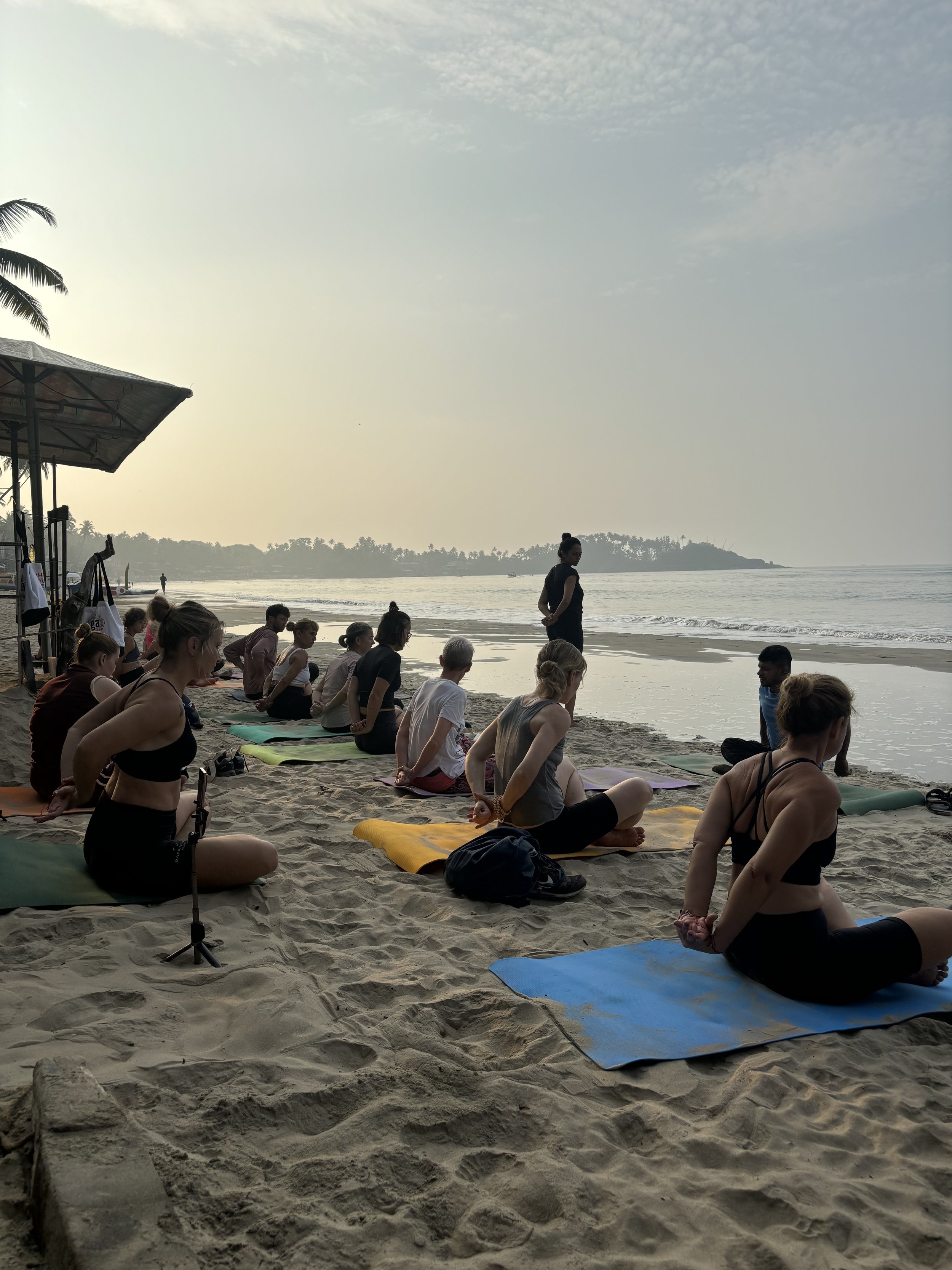Group yoga session on sandy beach at sunrise in Goa India, people seated on mats, ocean and distant palm trees in background.