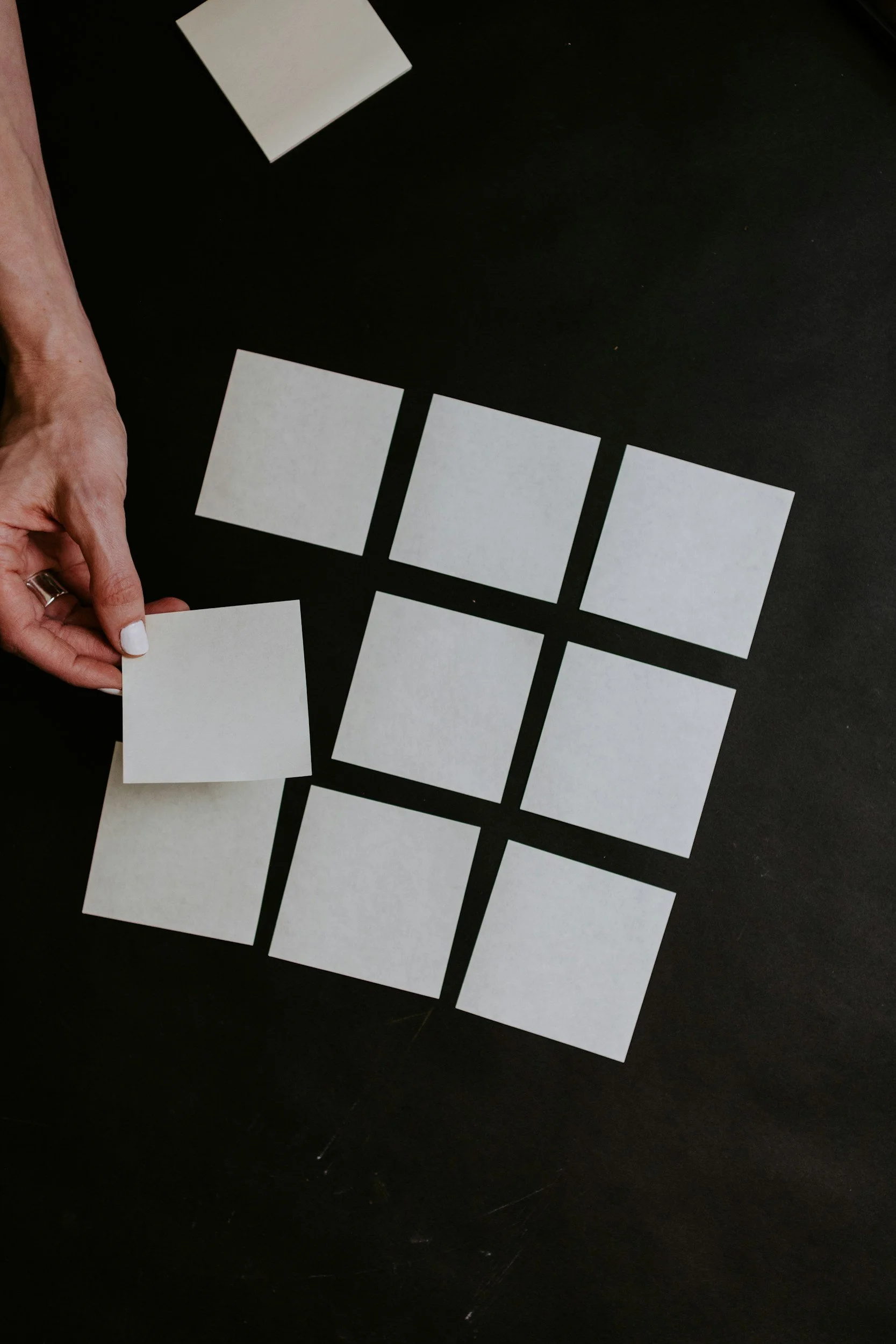 A person is holding a blank white square card over a black table with other similar cards arranged in a grid pattern.