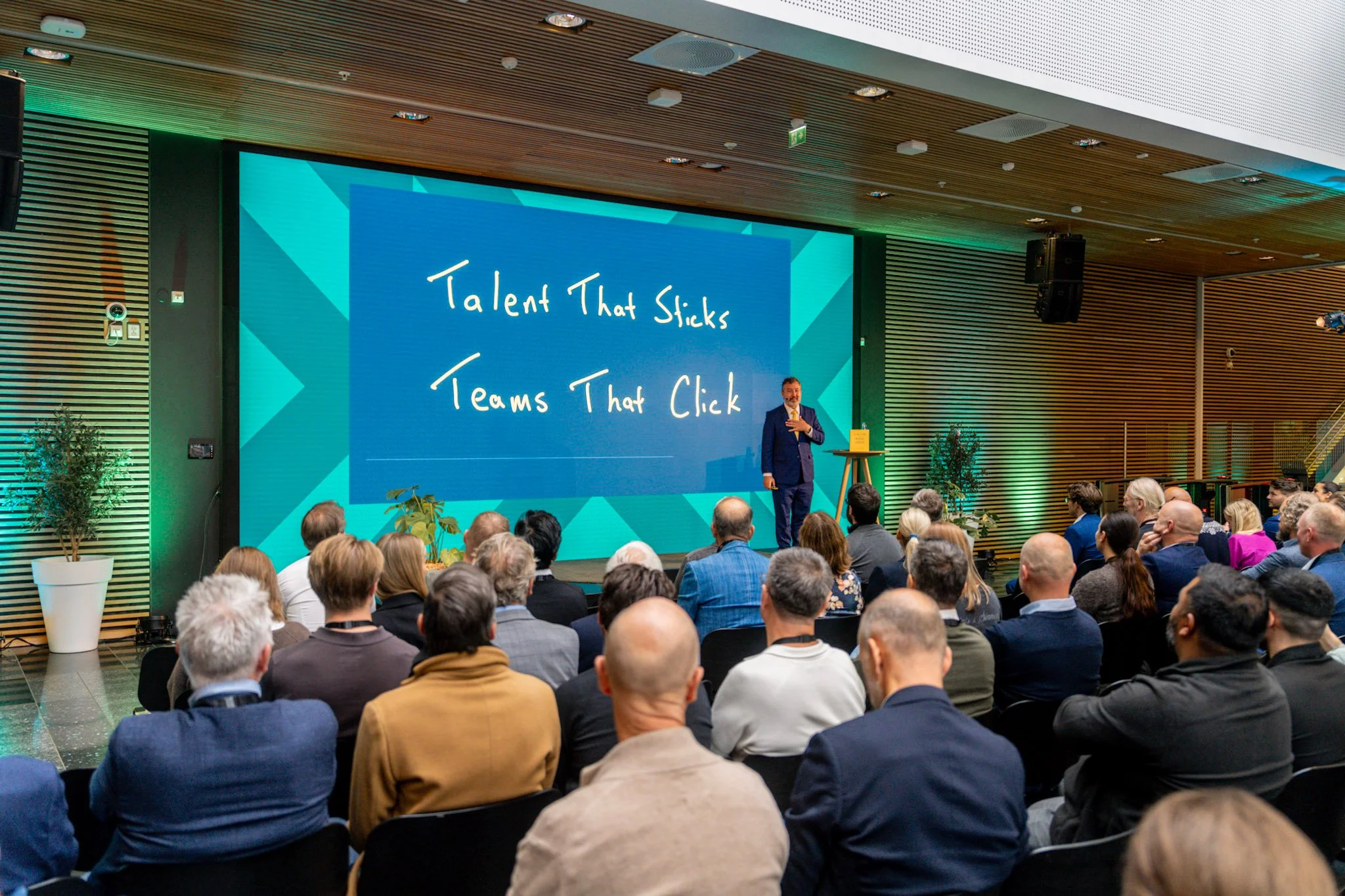 A speaker presenting to a full audience at a professional conference, with a large screen behind displaying the message “Talent That Sticks, Teams That Click.”